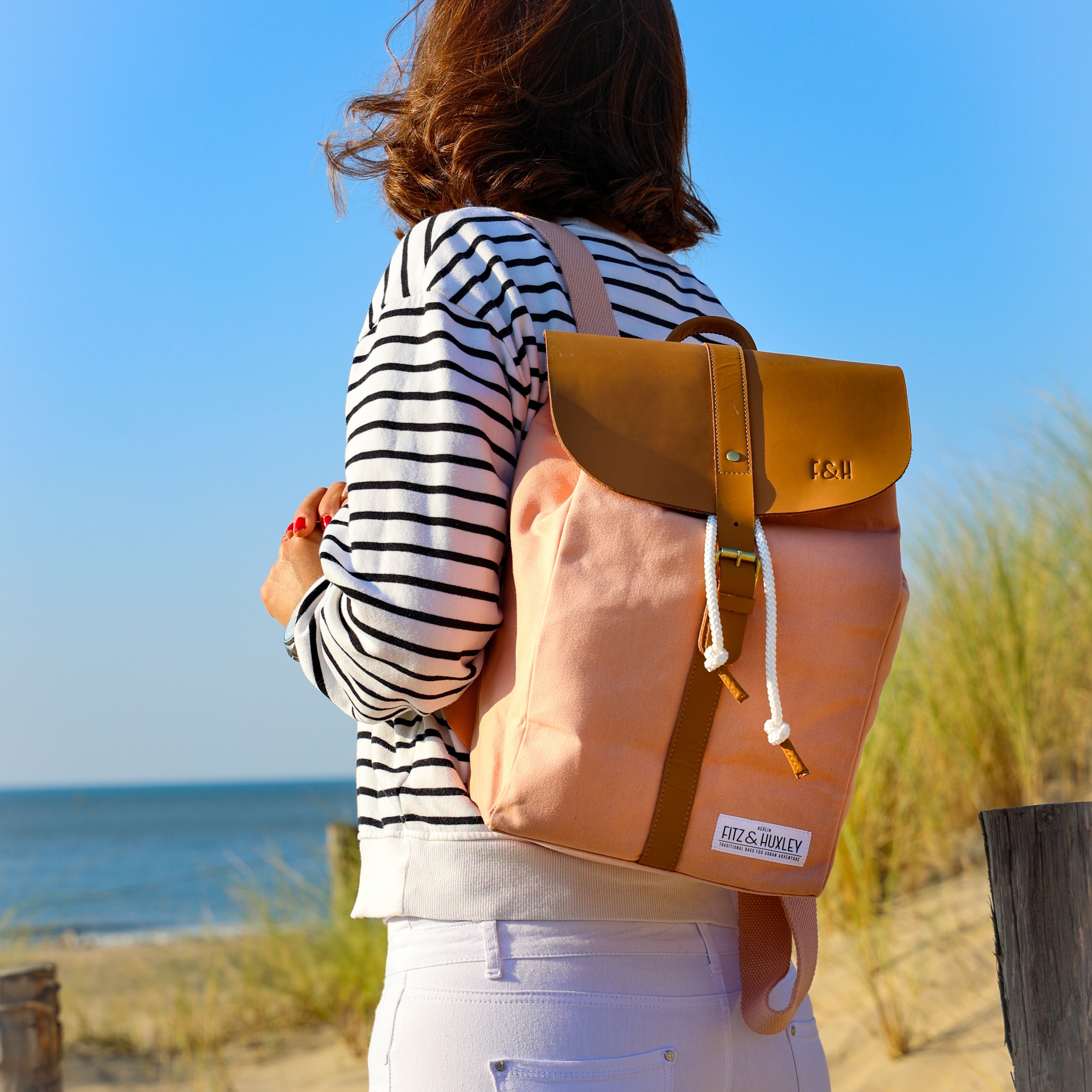 Frau in gestreiftem Pullover mit Rucksack blickt am Strand aufs Meer.