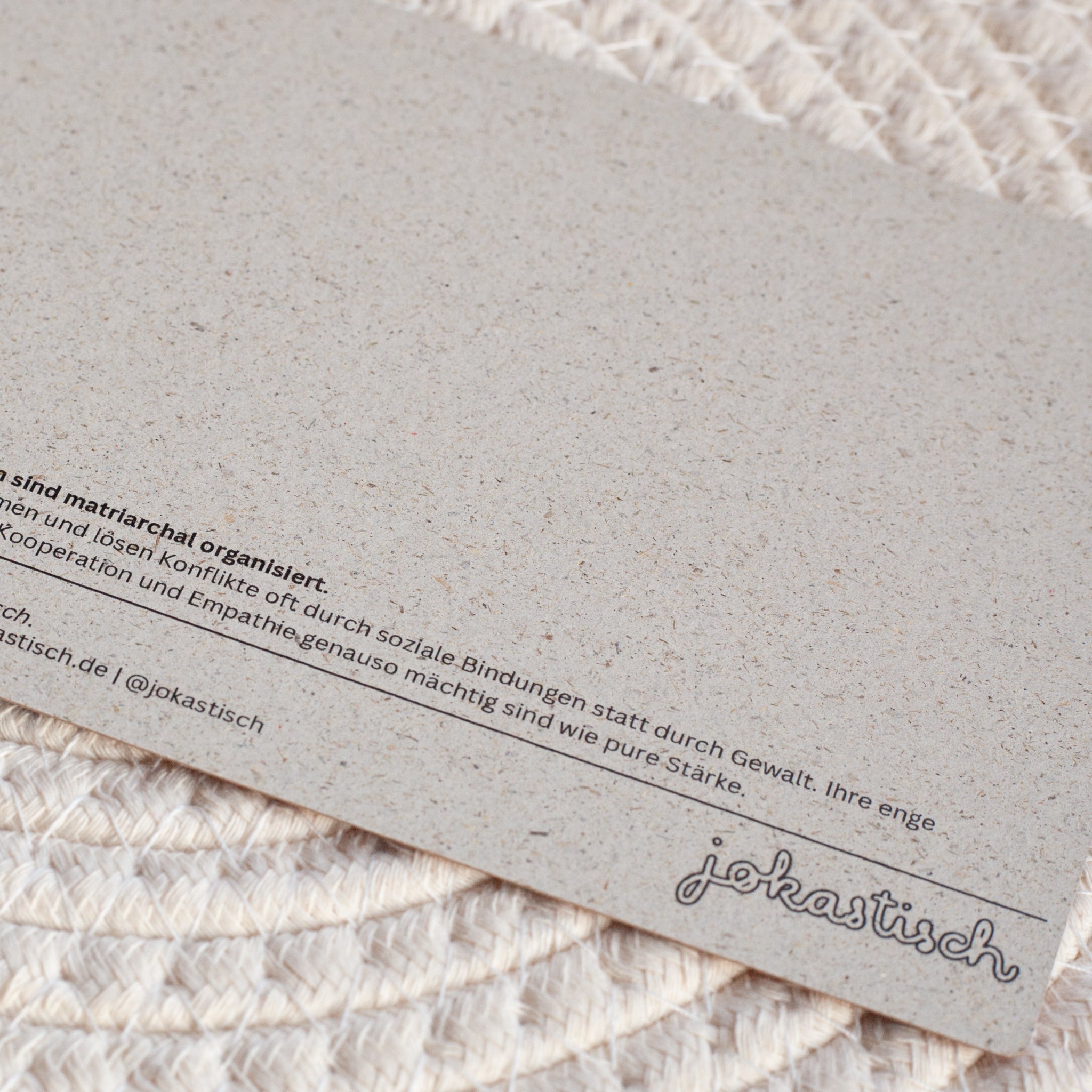 Nahaufnahme einer strukturierten Karte mit deutschem Text und dem Logo jokastisch in der rechten unteren Ecke, die auf einem gewebten, cremefarbenen Tischset liegt.