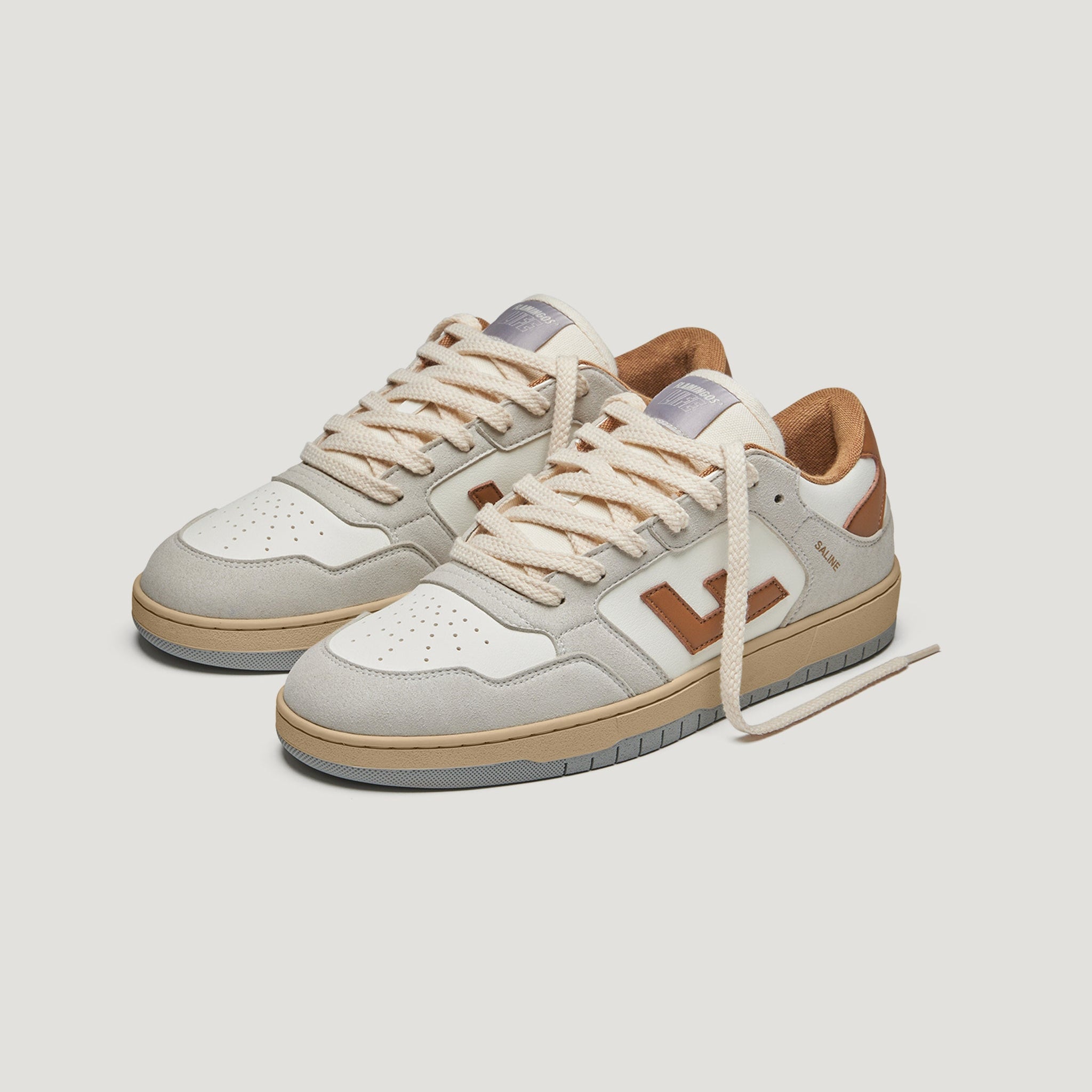 Ein Paar beige und hellgraue Sneaker mit hellbraunen Schnürsenkeln, hellbraunen und braunen Details und einer dicken Gummisohle. Die Schuhe haben Perforationen an der Spitze und ein braunes Logo an der Seite.