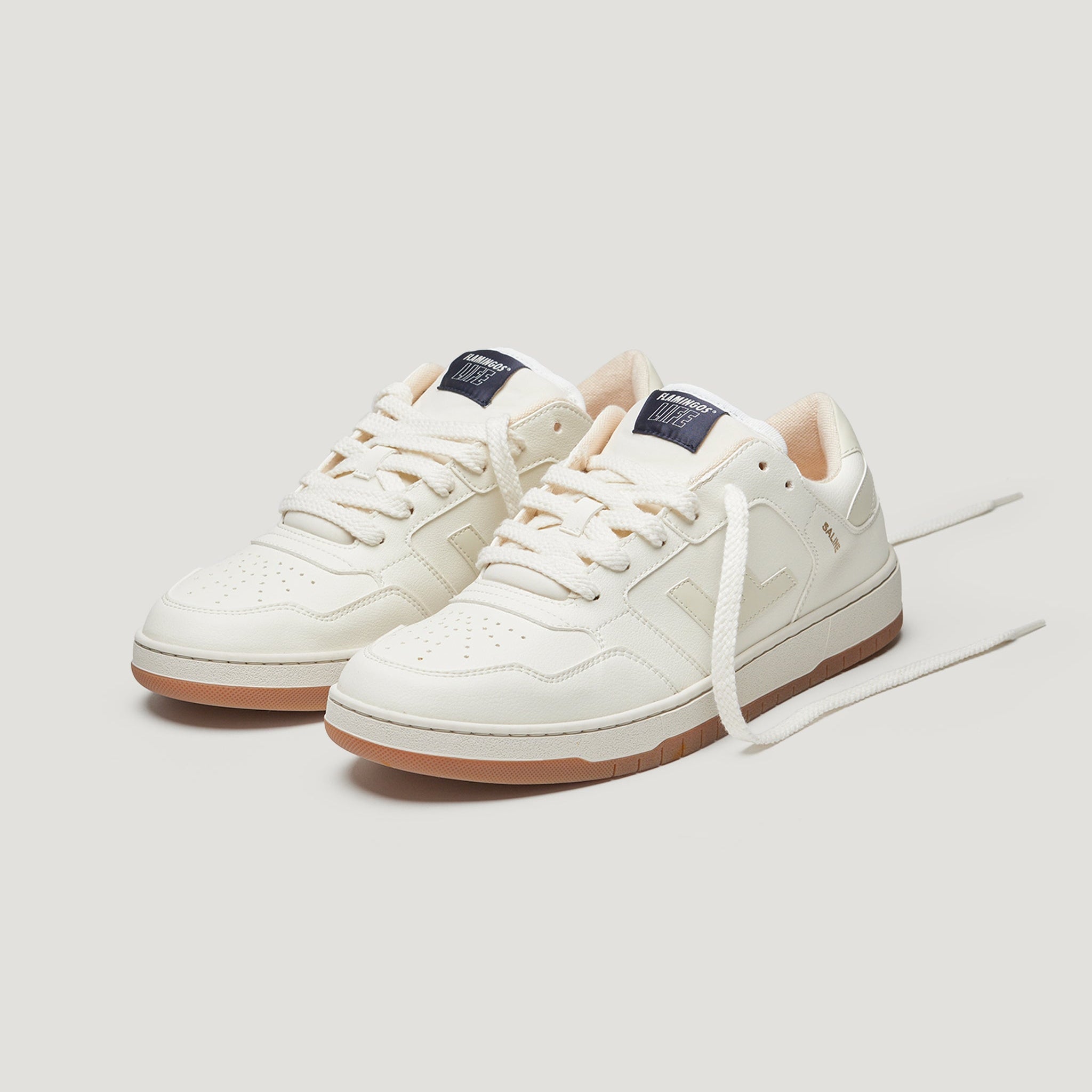 Ein Paar cremefarbene Turnschuhe mit weißen Schnürsenkeln und gummifarbenen Sohlen, mit einem marinefarbenen Aufnäher auf der Zunge, auf einem schlichten hellen Hintergrund.