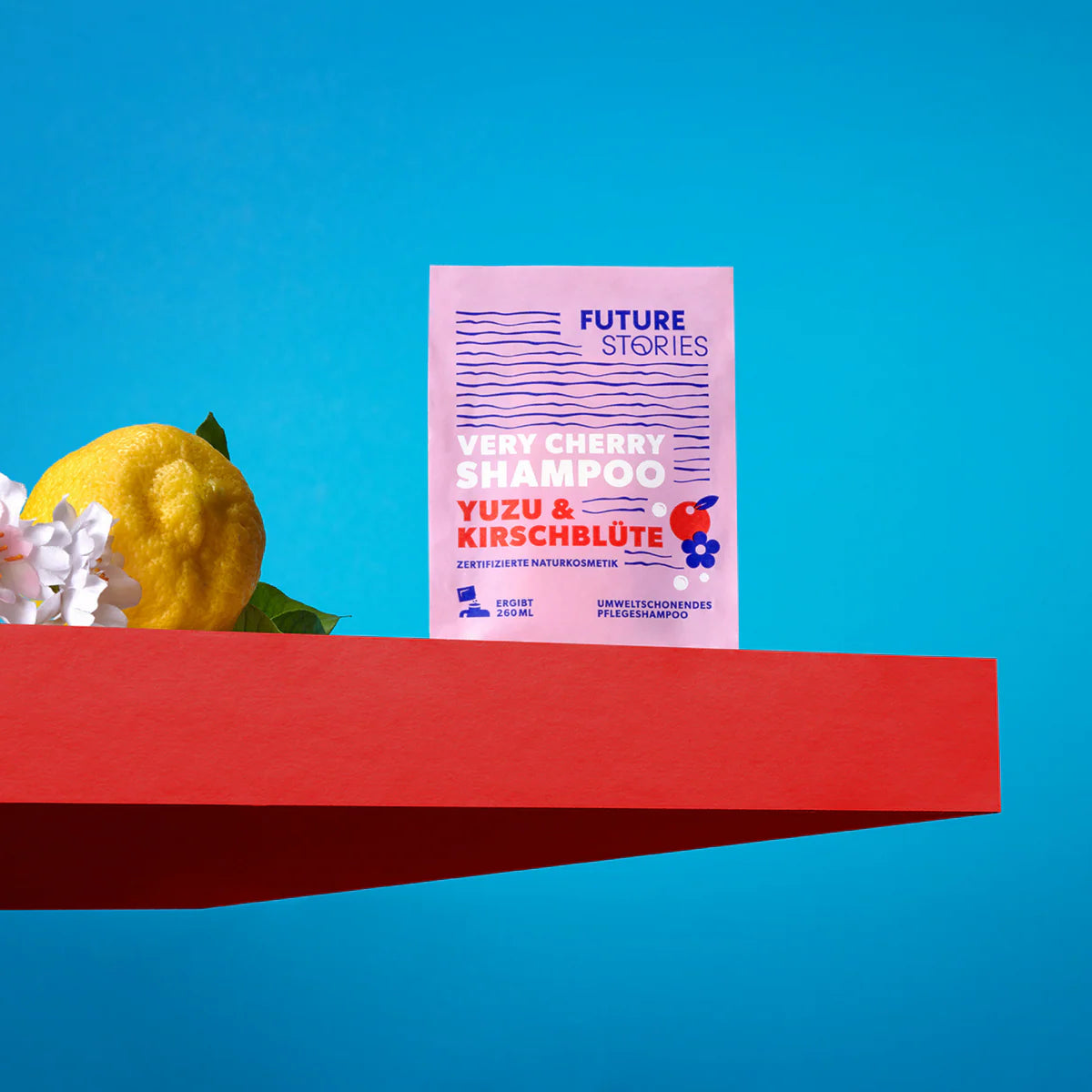 Ein Beutel Future Stories Very Cherry Shampoo liegt auf einer roten Fläche mit einer Yuzu-Frucht und weißen Blüten vor einem hellblauen Hintergrund. Die Verpackung ist rosa mit buntem Text und Illustrationen von Kirschen und Blumen.