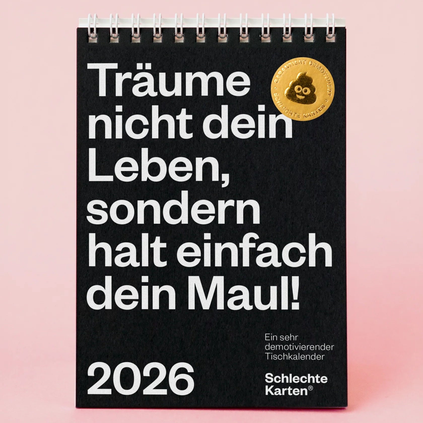 Ein schwarzer Spiral-Tischkalender mit fetter deutscher Schrift: Träume nicht dein Leben, sondern halt einfach dein Maul! 2026. Oben rechts befindet sich ein goldener Aufkleber. Darauf ist ein weißes Etikett: Jetzt neu: 52 Wochen. Rosa Hintergrund.