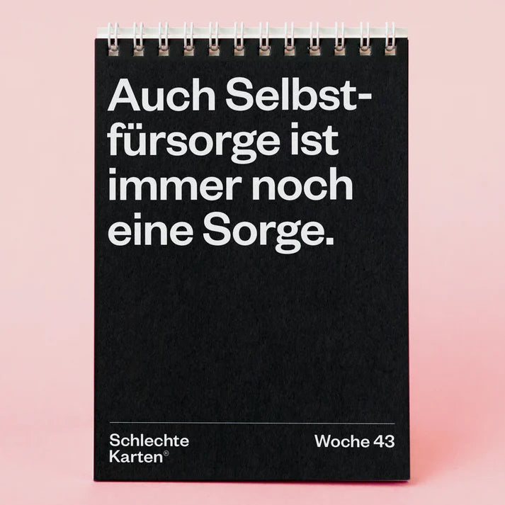 Ein schwarzer Spiral-Tischkalender auf rosa Hintergrund zeigt einen deutschen Text: Auch Selbstfürsorge ist immer noch eine Sorge. Unten stehen in weißen Buchstaben Schlechte Karten® und Woche 43.