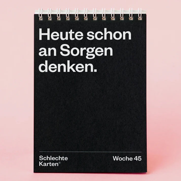 Ein schwarzer Spiral-Klappkalender steht auf einer rosa Fläche. Auf der Vorderseite steht in weißer Schrift "Heute schon an Sorgen denken.", am unteren Rand sind "Schlechte Karten" und "Woche 45" aufgedruckt.
