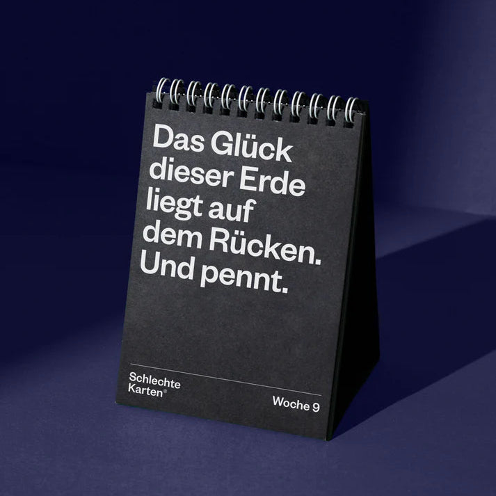Ein schwarzer Spiral-Tischkalender mit weißem deutschen Text, der lautet: Das Glück dieser Erde liegt auf dem Rücken. Und pennt. Am unteren Rand stehen Schlechte Karten und Woche 9. Der Hintergrund ist dunkelblau.