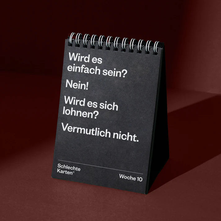 Ein schwarzer, spiralförmiger Tischkalender steht auf einer dunkelroten Fläche. Auf Deutsch steht darauf: Wird es einfach sein? Nein! Wird es sich lohnen? Vermutlich nicht. Unterer Text: Schlechte Karten® Woche 10.