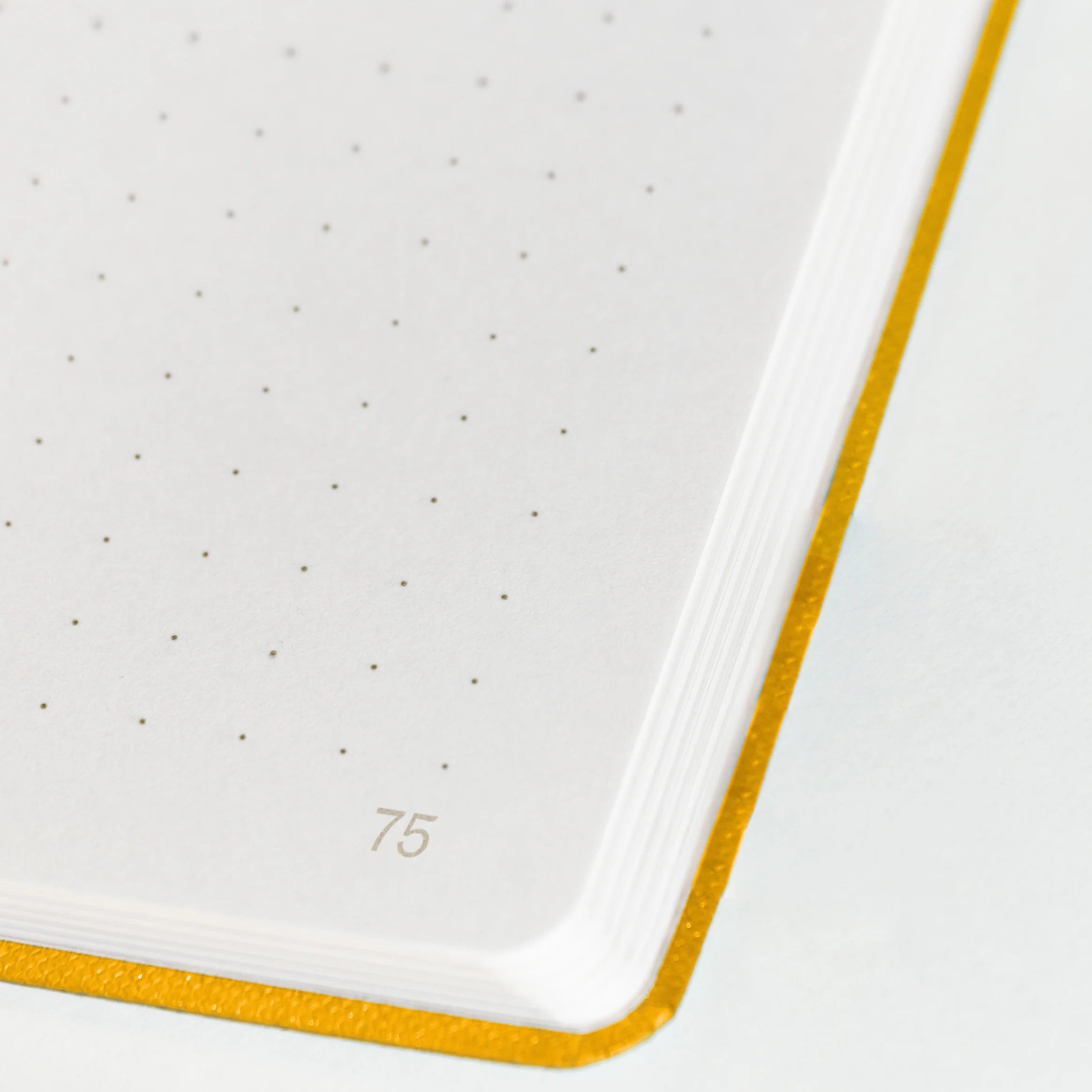 Nahaufnahme der rechten unteren Ecke eines aufgeschlagenen Notizbuchs mit gepunktetem Rasterpapier, einem gelben Einband und der Zahl 75 auf der Seite.