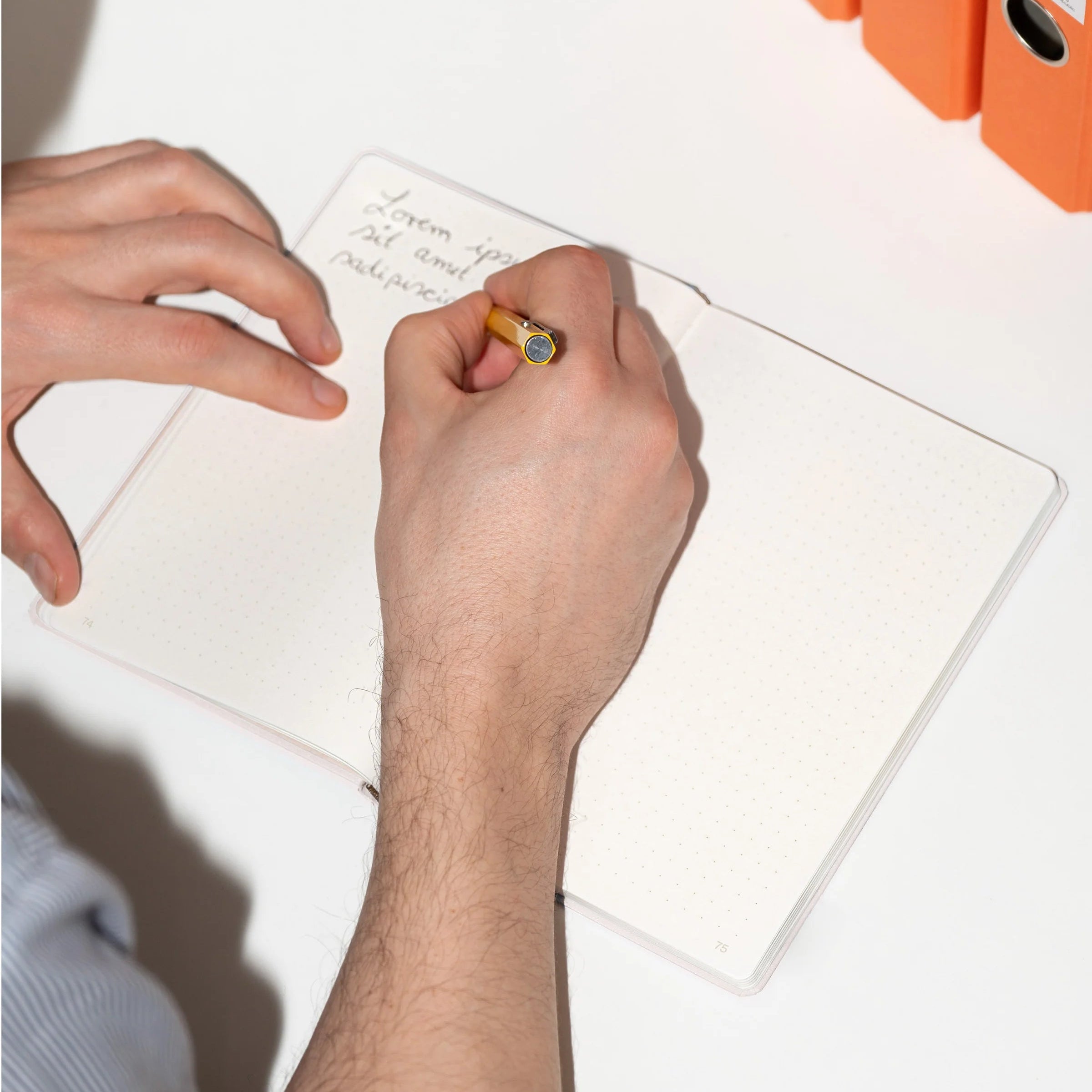 Eine Person schreibt an einem weißen Schreibtisch mit einem gelben Bleistift in ein gepunktetes Notizbuch. Drei orangefarbene Aktenordner sind in der Nähe gestapelt, und die Person trägt ein hellblaues Hemd.