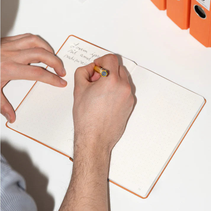 Eine Person schreibt in einem gepunkteten Notizbuch mit einem Bleistift an einem weißen Schreibtisch. In der Ecke sind drei orangefarbene Aktenordner zu sehen. Der Text im Notizbuch ist teilweise sichtbar und handgeschrieben.