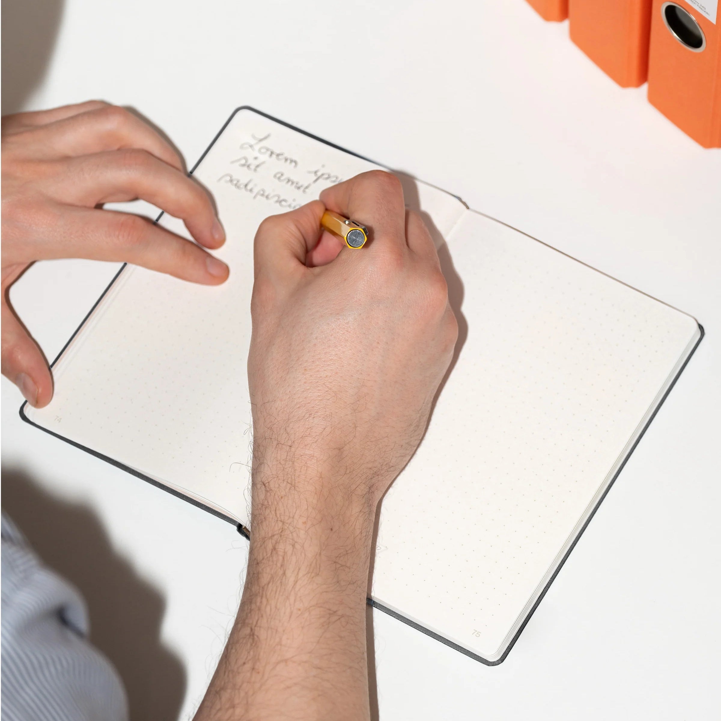Eine Person schreibt mit einem Bleistift in einem gepunkteten Notizbuch auf einem weißen Schreibtisch. In der Nähe sind drei orangefarbene Ringbücher gestapelt. Die Schrift in dem Notizbuch ist teilweise sichtbar.