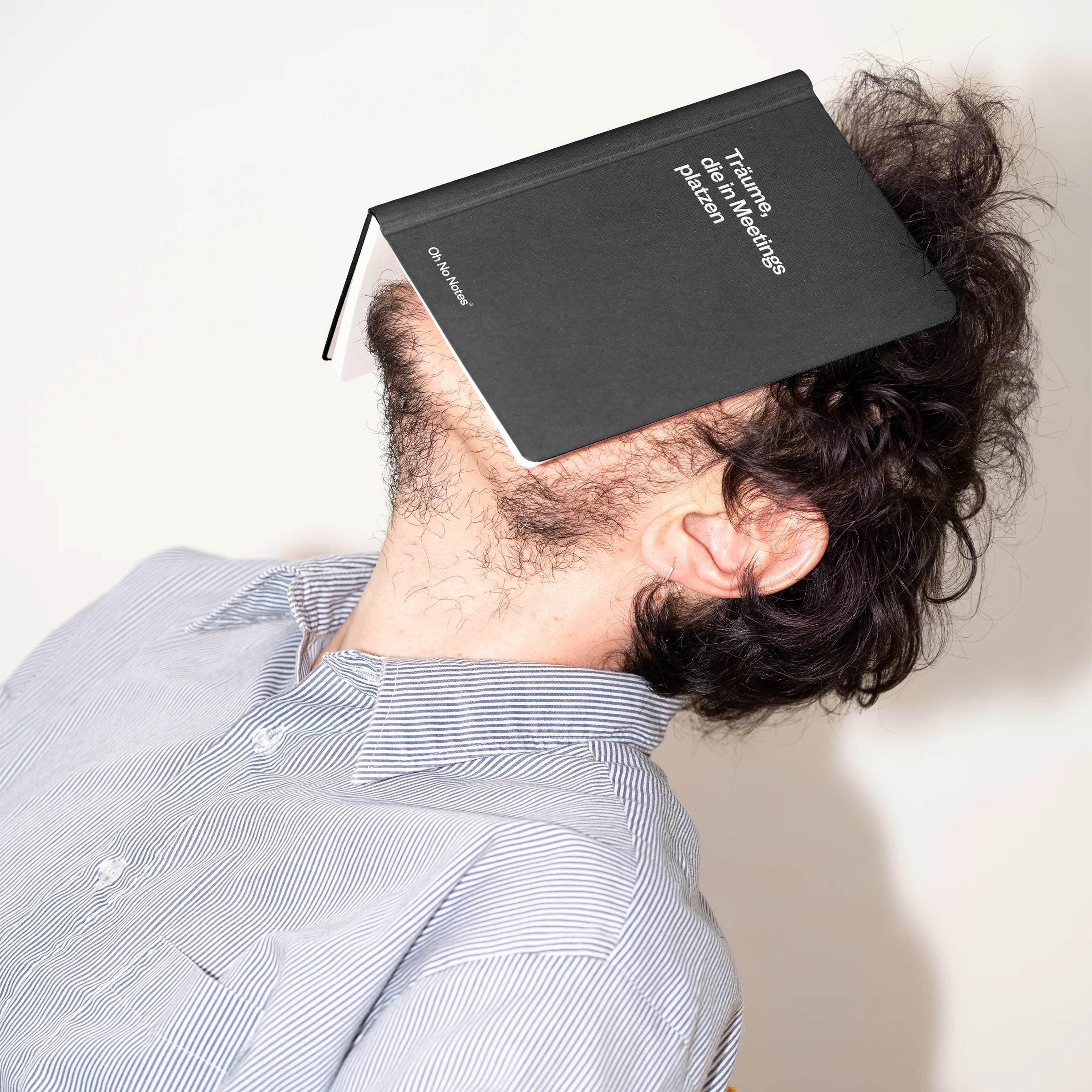 Eine Person mit zerzaustem Haar lehnt sich in einem Stuhl zurück, ein geschlossenes schwarzes Buch liegt mit dem Gesicht nach unten über ihrem Gesicht, so dass ihre Gesichtszüge verdeckt sind. Sie trägt ein gestreiftes Hemd vor einem schlichten Hintergrund.