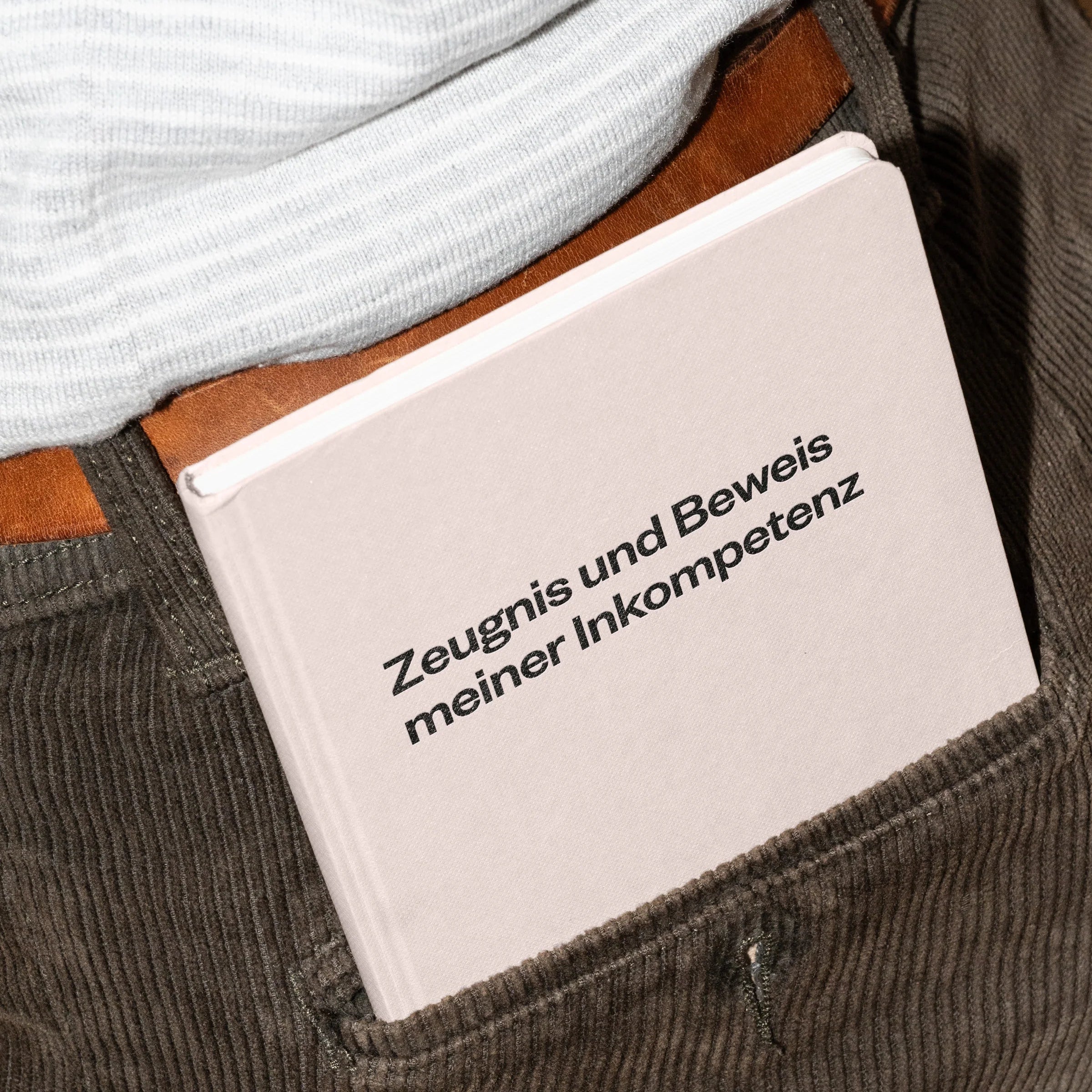 Ein hellrosa Buch mit dem deutschen Text Zeugnis und Beweis meiner Inkompetenz steckt in der Gesäßtasche einer braunen Cordhose. Ein weißes Hemd ist teilweise über dem Buch zu sehen.