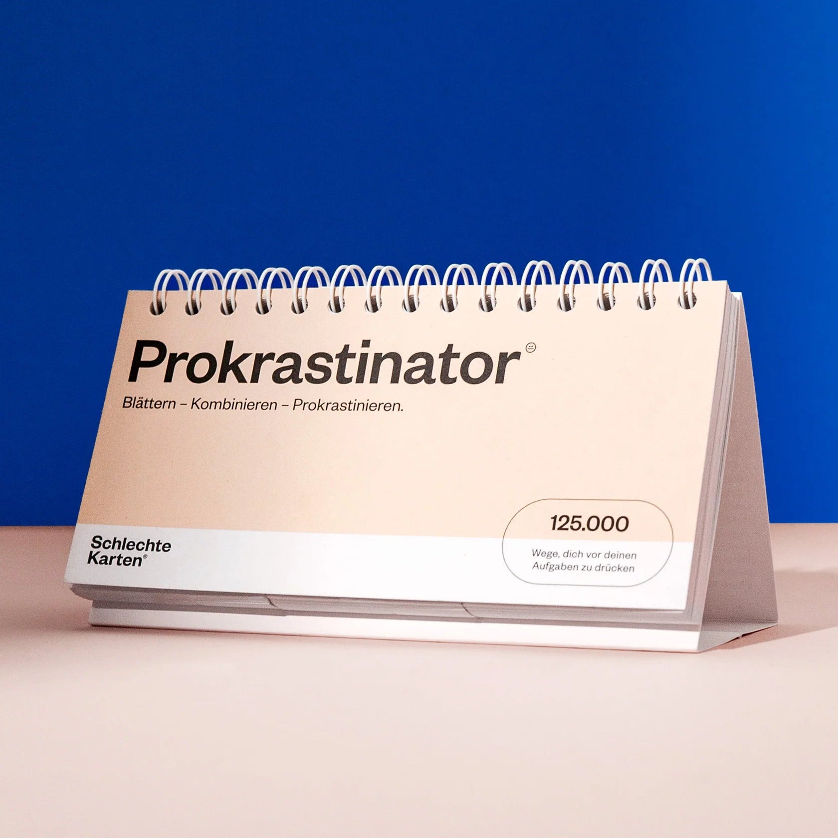 Ein spiralgebundener Tischkalender mit dem Titel Prokrastinator steht auf einer rosa Fläche vor einem blauen Hintergrund. Der Einband des Kalenders ist beige und weist einen deutschen Text und ein rundes Abzeichen mit der Aufschrift 125.000 auf.