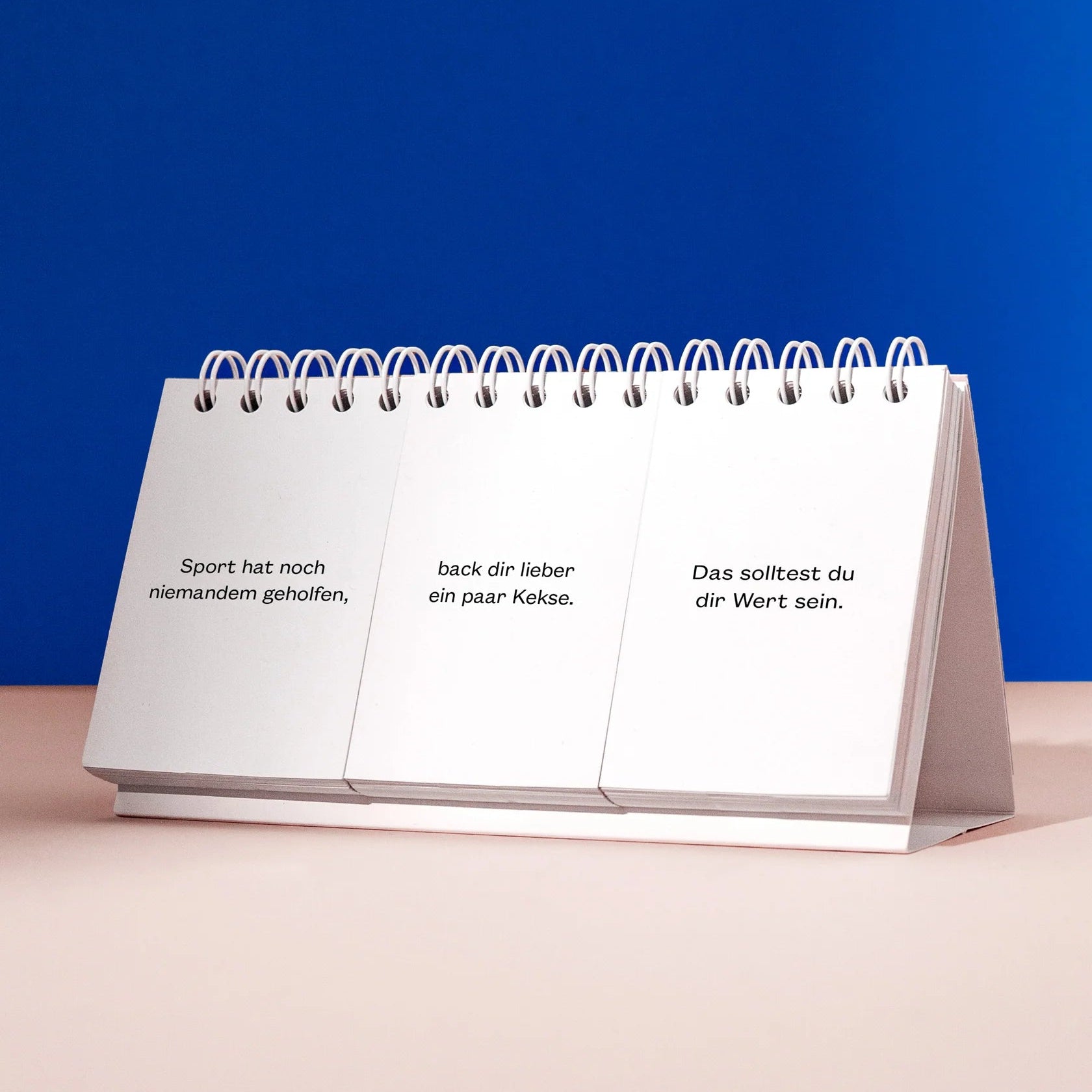 Ein weißer Klappkalender mit deutschem Text auf drei Seiten vor blauem Hintergrund auf einer hellrosa Oberfläche. Der Text lautet: Sport hat noch niemandem geholfen, back dir lieber ein paar Kekse. Das solltest du dir Wert sein.