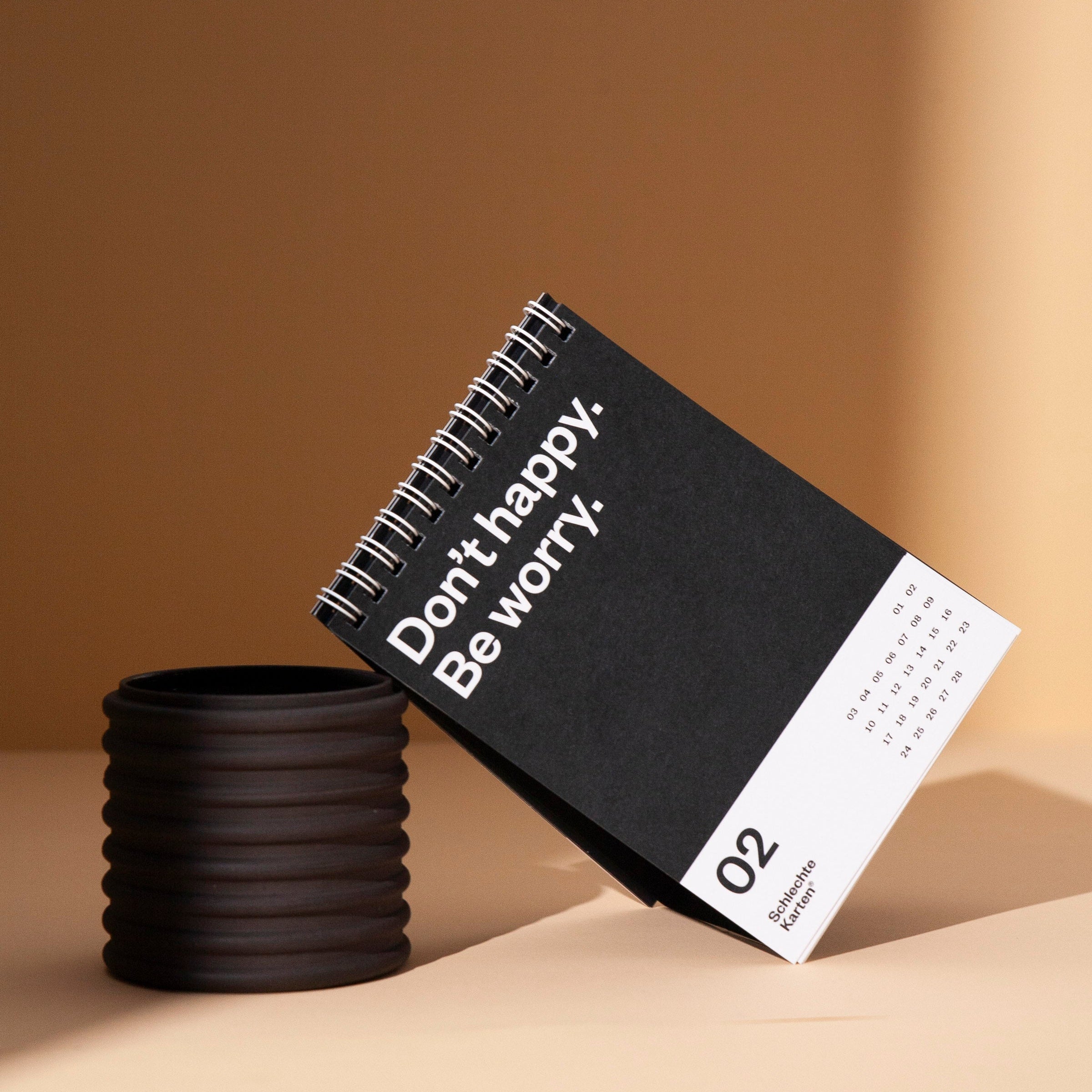 Kalender mit spiralgebundener Karte zeigt "Don't happy. Be worry." über einem dunklen Behälter auf beigem Hintergrund.
