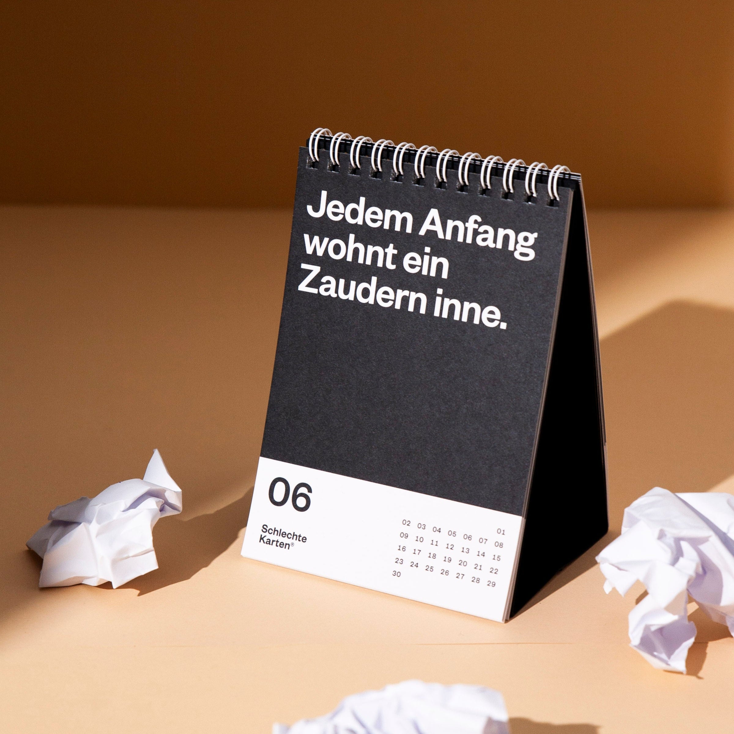 Ein schwarzer Tischkalender mit dem Zitat "Jedem Anfang wohnt ein Zaudern inne" und zerknülltem Papier daneben.