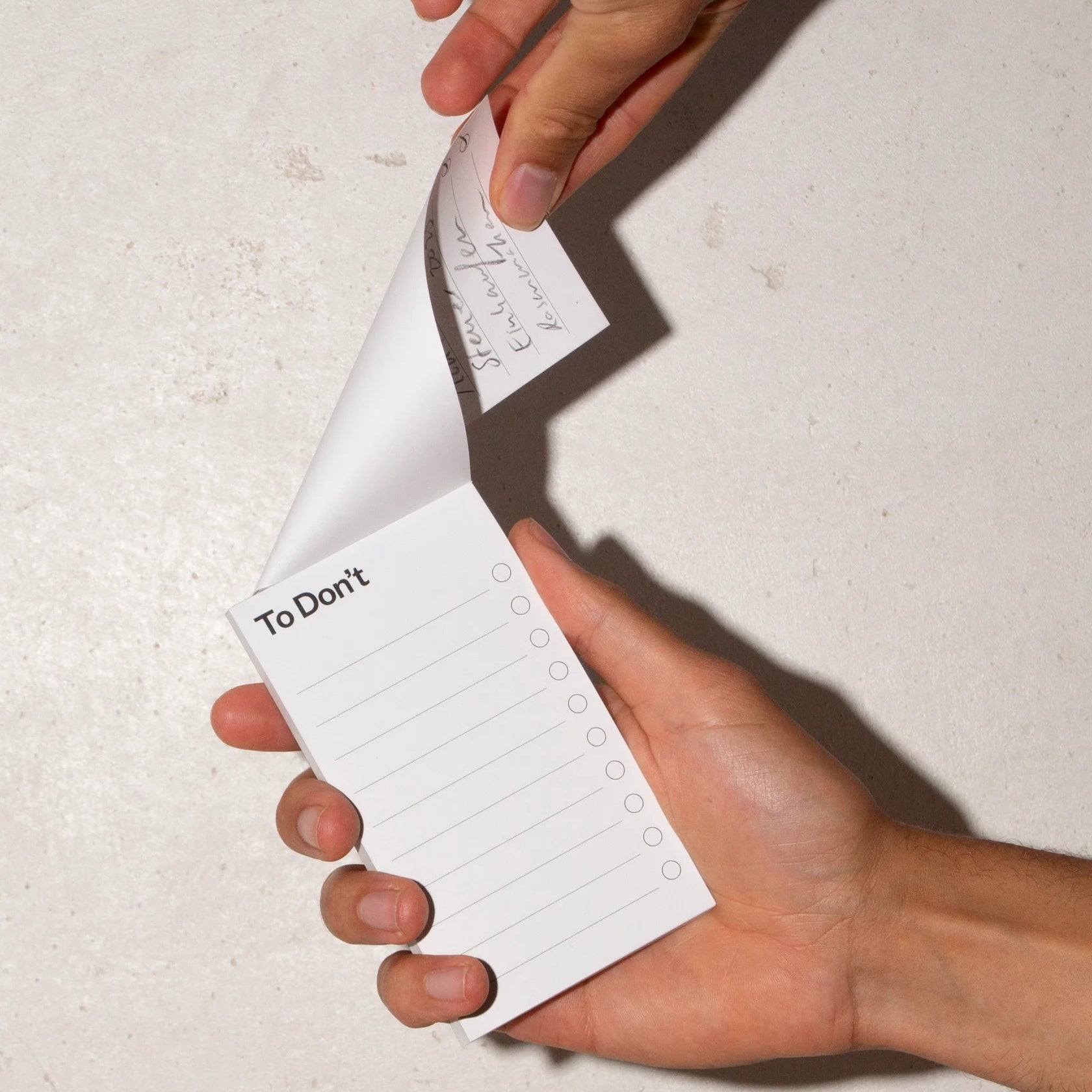 Zwei Hände halten einen Notizblock mit der Aufschrift To Don't und leeren Kästchen. Eine Hand blättert eine Seite um, um handschriftliche Notizen auf dem darunter liegenden Blatt sichtbar zu machen. Der Hintergrund ist eine glatte, strukturierte Oberfläche.
