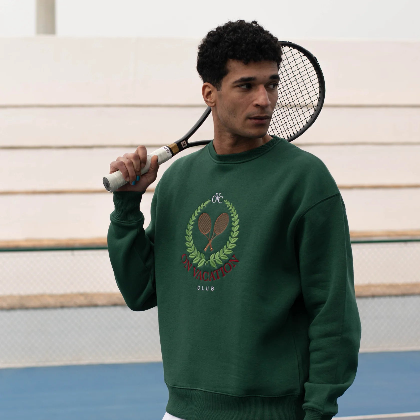 Ein Mann in einem grünen Pullover mit Tennisschläger steht vor leeren Tribünen.