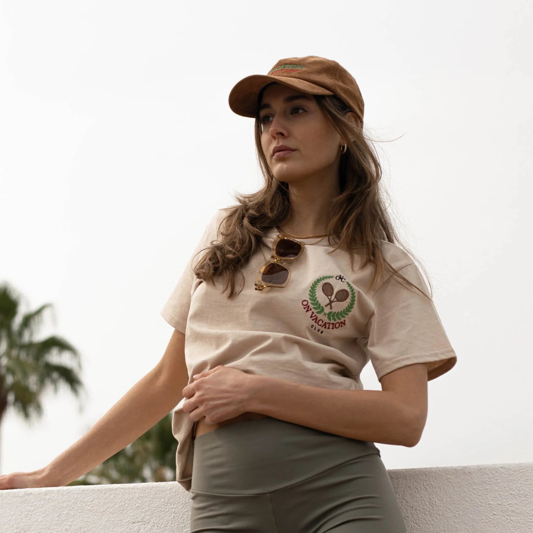 Frau mit braunem Hut und beigem "On Vacation"-Shirt steht entspannt draußen, Sonnebrille in der Mitte des Shirts.