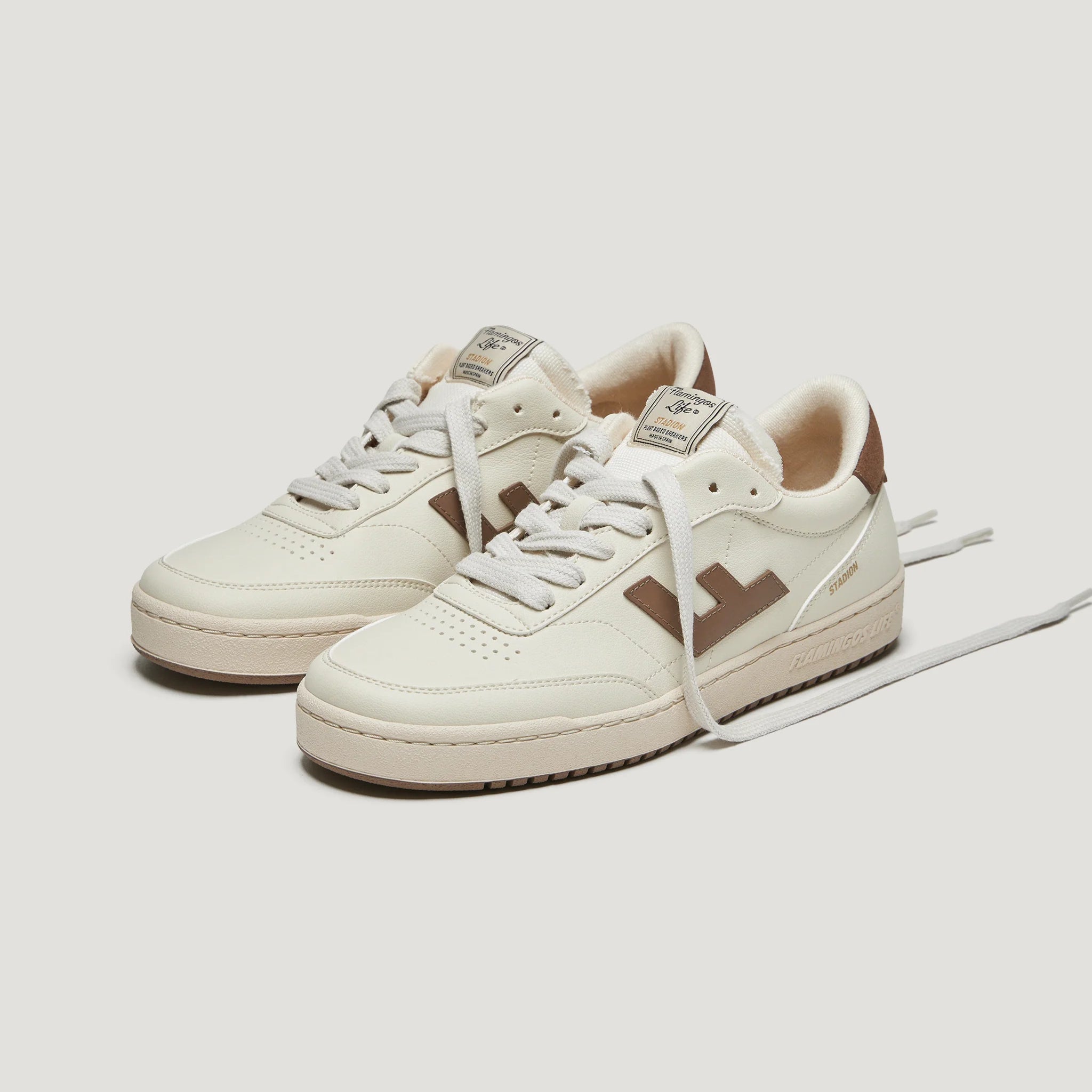 Weiße Sportschuhe mit braunen Details und weißen Schnürsenkeln auf hellgrauem Hintergrund.