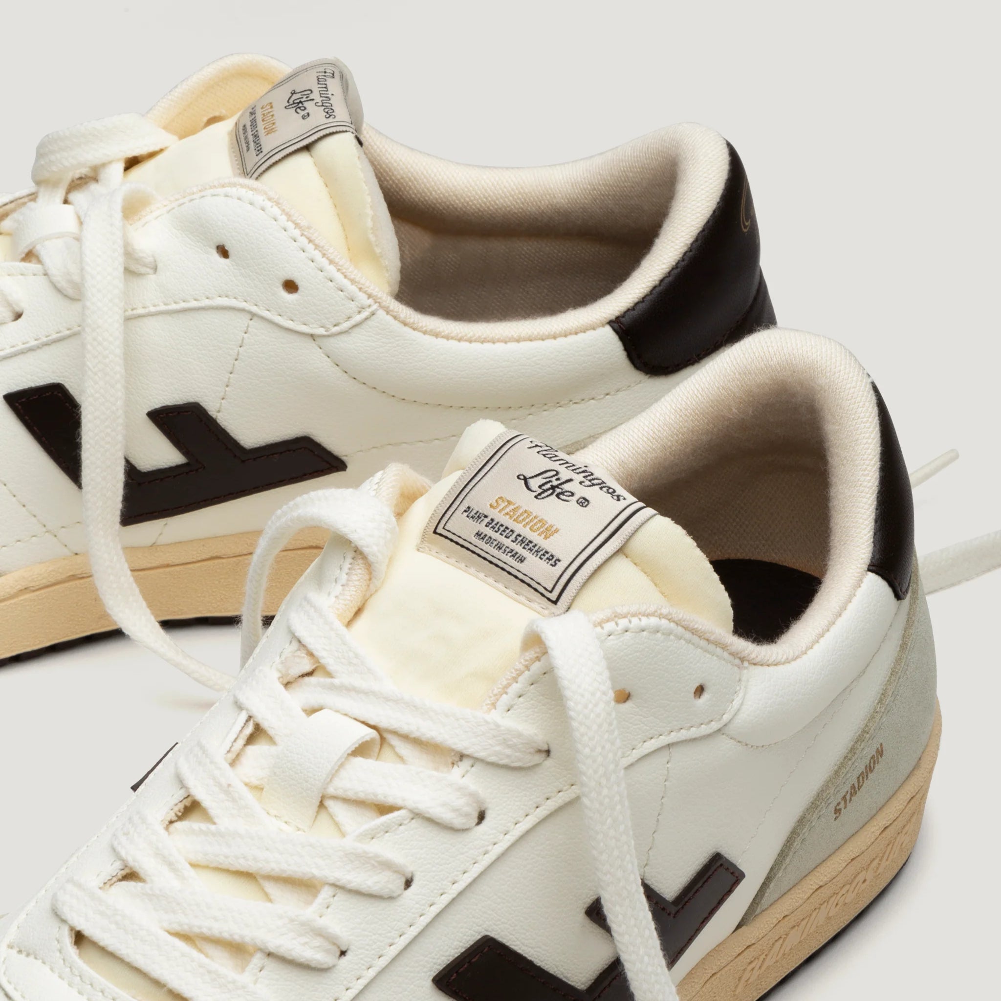 Weiße Sneaker mit braunen Details, cremefarbenen Schnürsenkeln und Etikett auf der Zunge, auf hellem Hintergrund.
