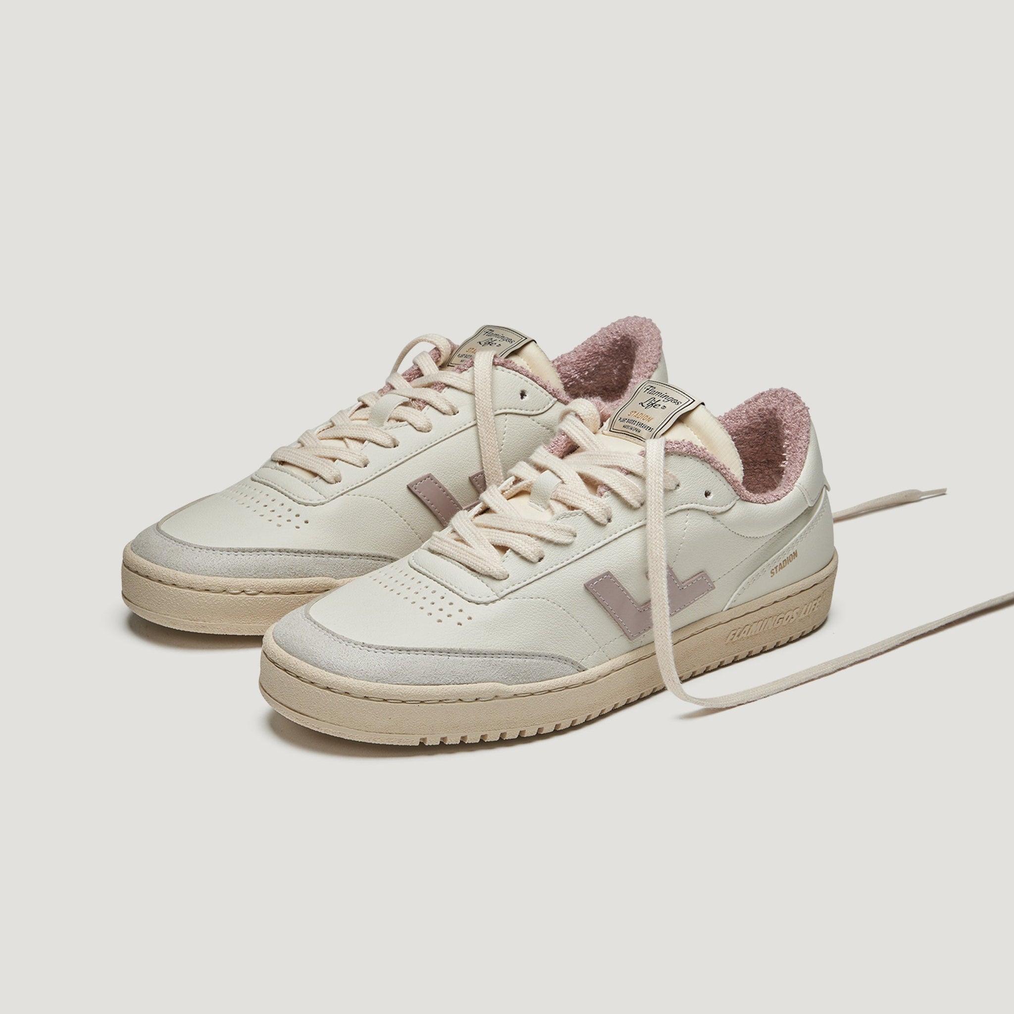 Ein Paar cremefarbene Turnschuhe mit rosa Akzenten, einem strukturierten rosa Futter und beigen Sohlen. Die Schuhe haben Schnürsenkel und ein auffälliges rosa L-Logo an der Seite.