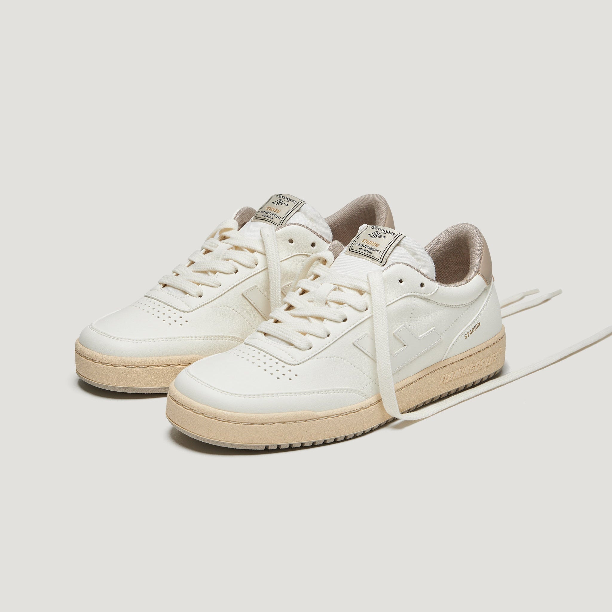 Ein Paar cremefarbene Ledersneaker mit beiger Sohle, dezenten Nähten und Branding an der Seite und auf der Zunge, dargestellt auf einem schlichten weißen Hintergrund. Ein Schnürsenkel ist locker auf der Oberfläche verlängert.