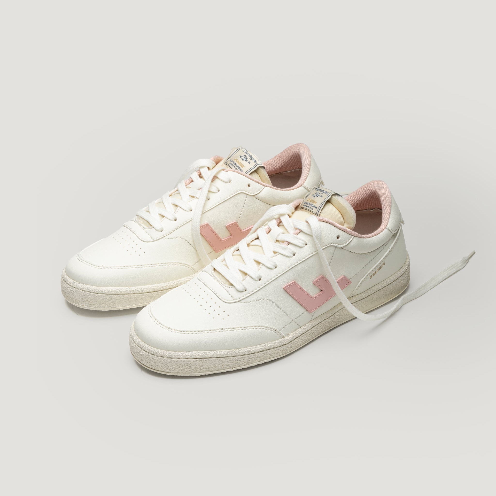 Ein Paar weiße Turnschuhe mit hellrosa Akzenten, einschließlich des Innenfutters und eines großen L-Logos an der Seite, vor einem schlichten hellen Hintergrund. Bei einem Schuh sind die Schnürsenkel teilweise geöffnet.