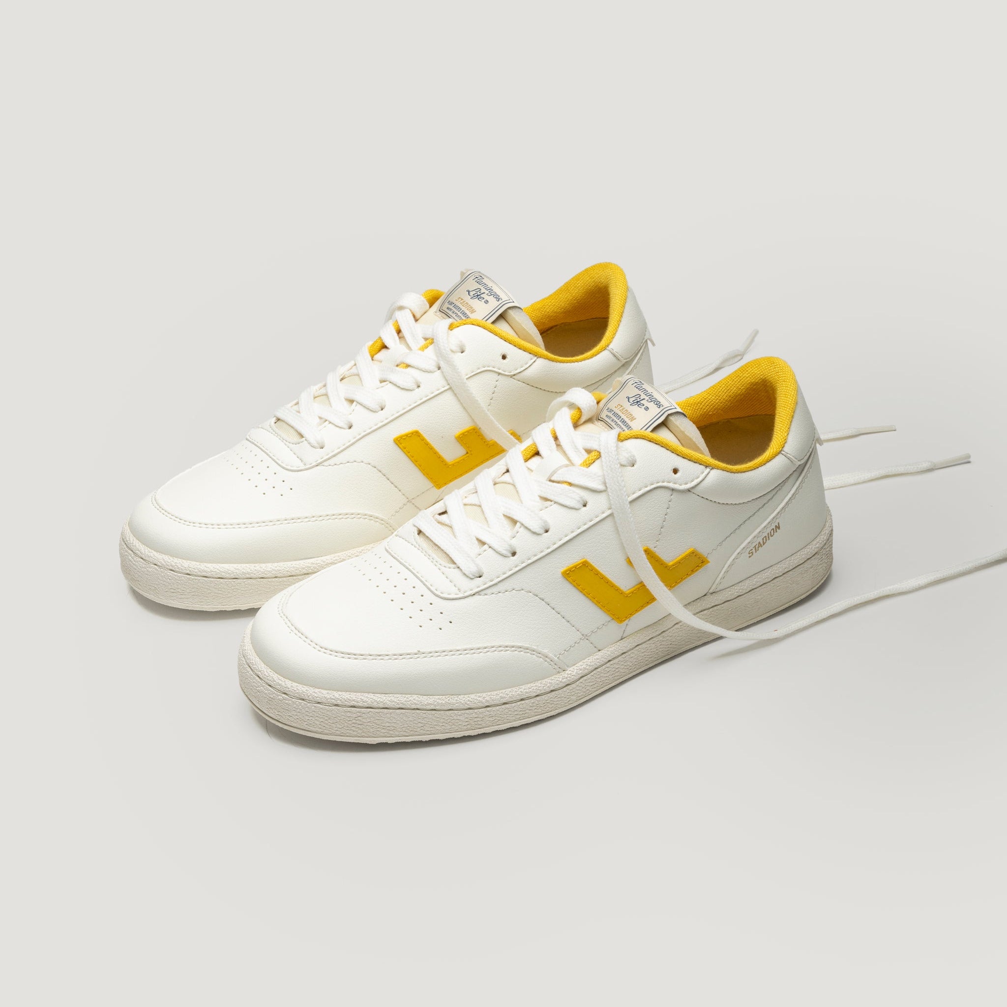 Ein Paar weiße Turnschuhe mit gelben Akzenten, einschließlich gelber L-Logos an den Seiten und gelbem Innenfutter, sind nebeneinander auf einem hellgrauen Hintergrund platziert. Die Schnürsenkel sind locker gebunden.