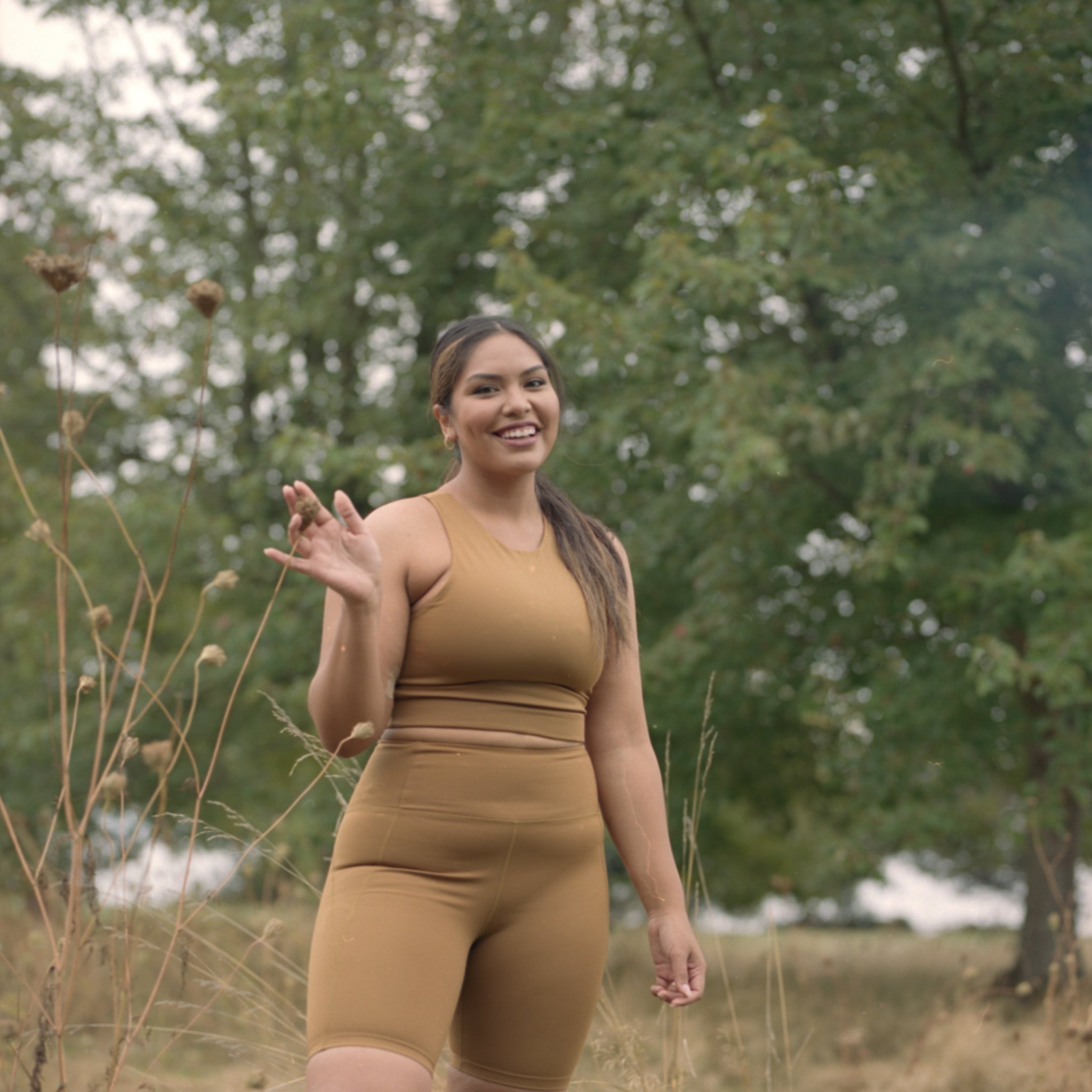 Person mit braunem Sportoutfit winkt und steht im Freien in einer grünen, bewaldeten Umgebung.