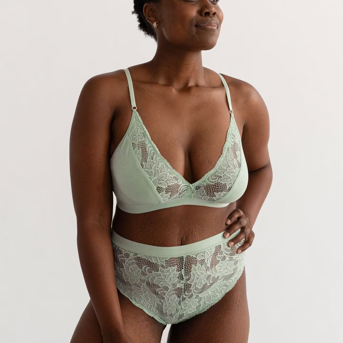 Frau trägt hellgrüne Spitzenunterwäsche, steht seitlich und lächelt selbstbewusst vor neutralem Hintergrund.