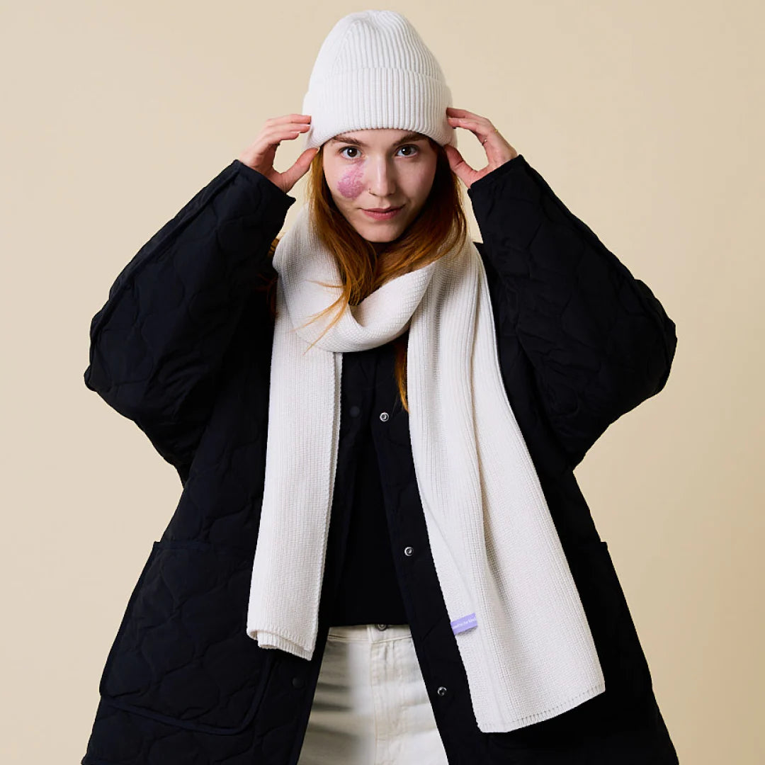Eine Frau mit schwarzem Steppmantel, weißem Strickschal und weißer Mütze posiert vor einem beigen Hintergrund und hält mit beiden Händen die Spitze ihres Hutes fest. Sie hat langes kastanienbraunes Haar und einen schwachen rosa Fleck auf der Wange.