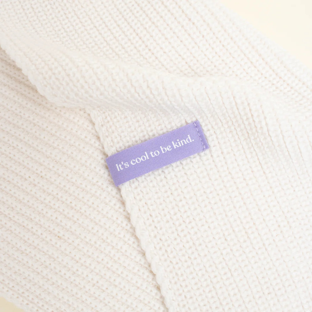Nahaufnahme eines weißen Strickstoffs mit einem aufgenähten lila Etikett mit der Aufschrift Its cool to be kind.