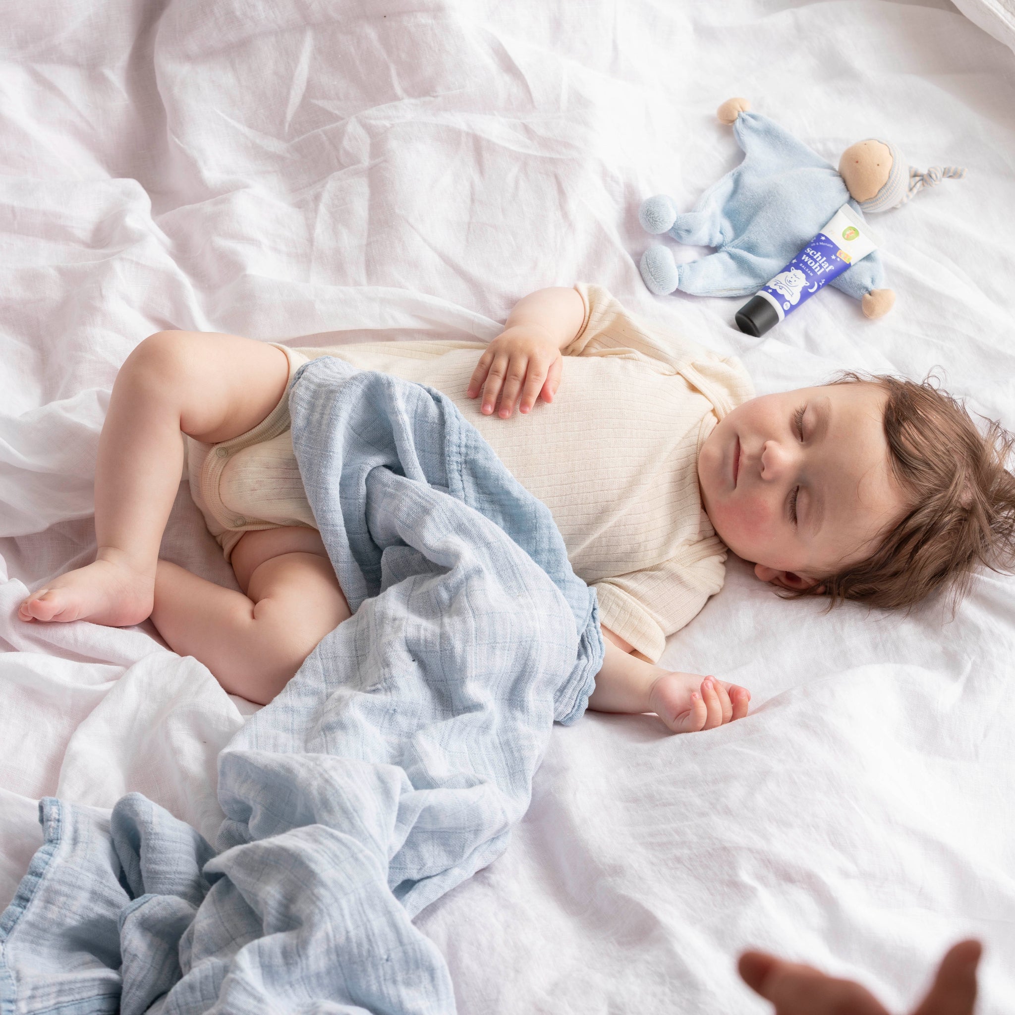 Ein Baby mit braunem Haar schläft friedlich in einem weißen Bett, das teilweise mit einer hellblauen Decke zugedeckt ist. In der Nähe befinden sich ein blaues Plüschtier und eine Tube mit Creme oder Salbe.