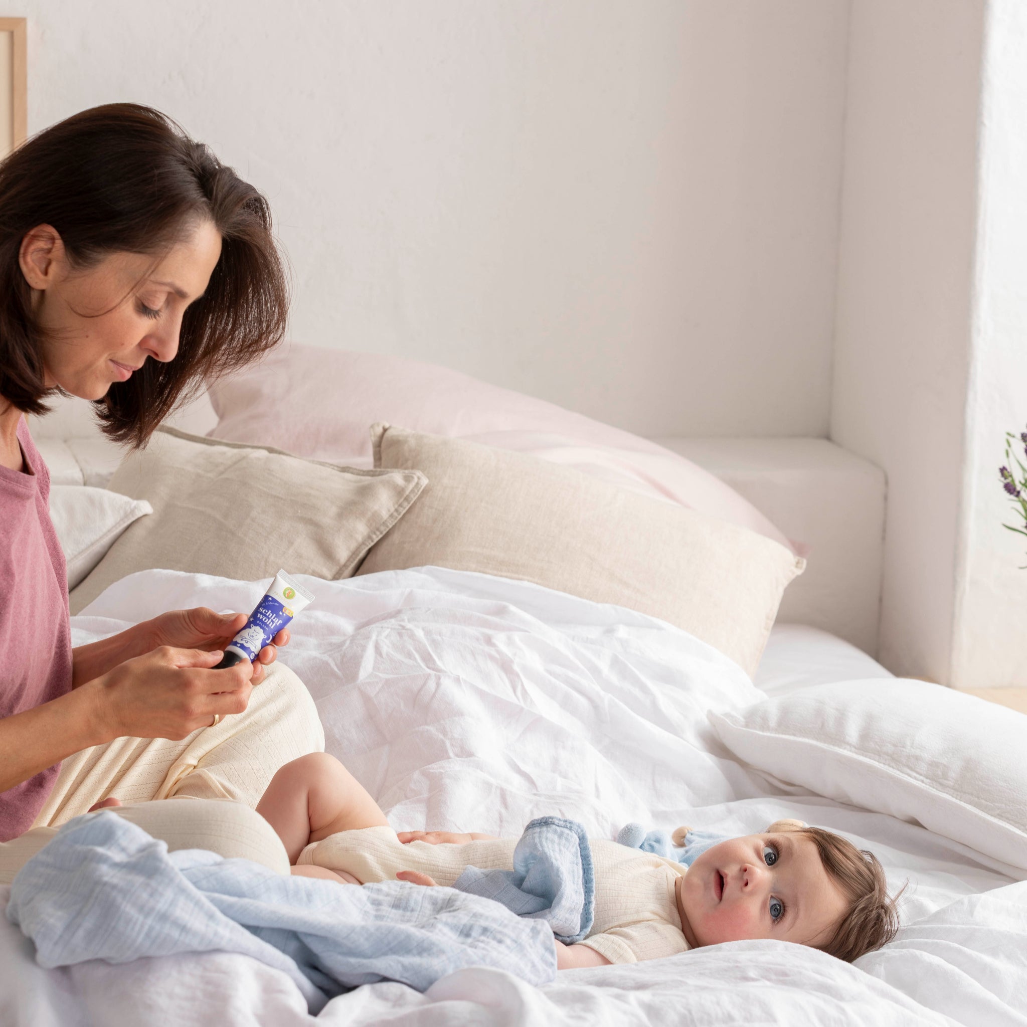Eine Frau sitzt auf einem Bett und hält eine Cremetube in der Hand, während ein Baby auf dem Bett liegt und nach oben schaut, teilweise zugedeckt mit einer hellblauen Decke. Der Raum ist sanft beleuchtet, Bettzeug und Kissen sind in neutralen Farben gehalten.
