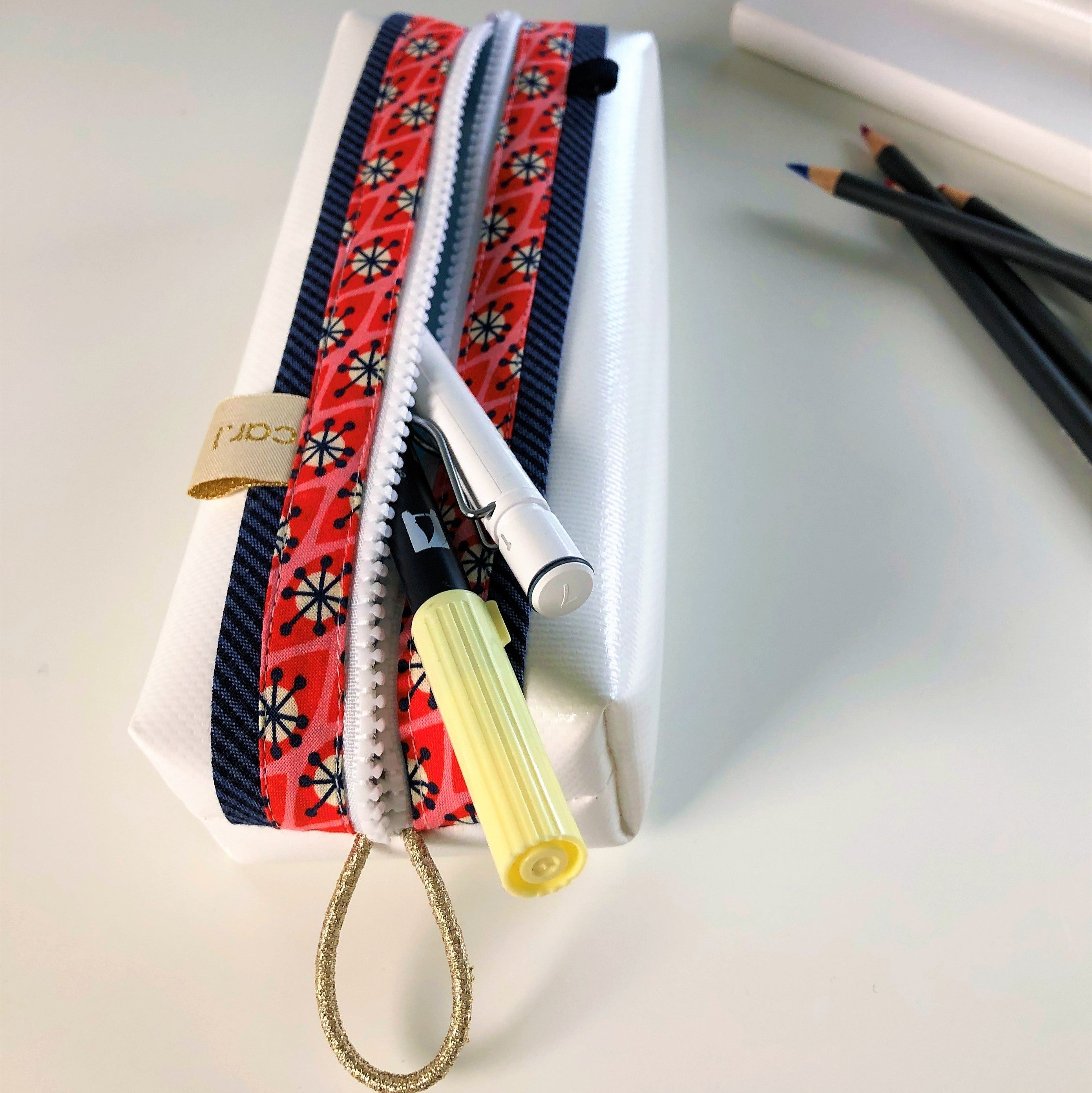 Ein Federmäppchen mit rotem Muster, das Stifte und einen gelben Marker enthält, neben einer Reihe von Buntstiften.