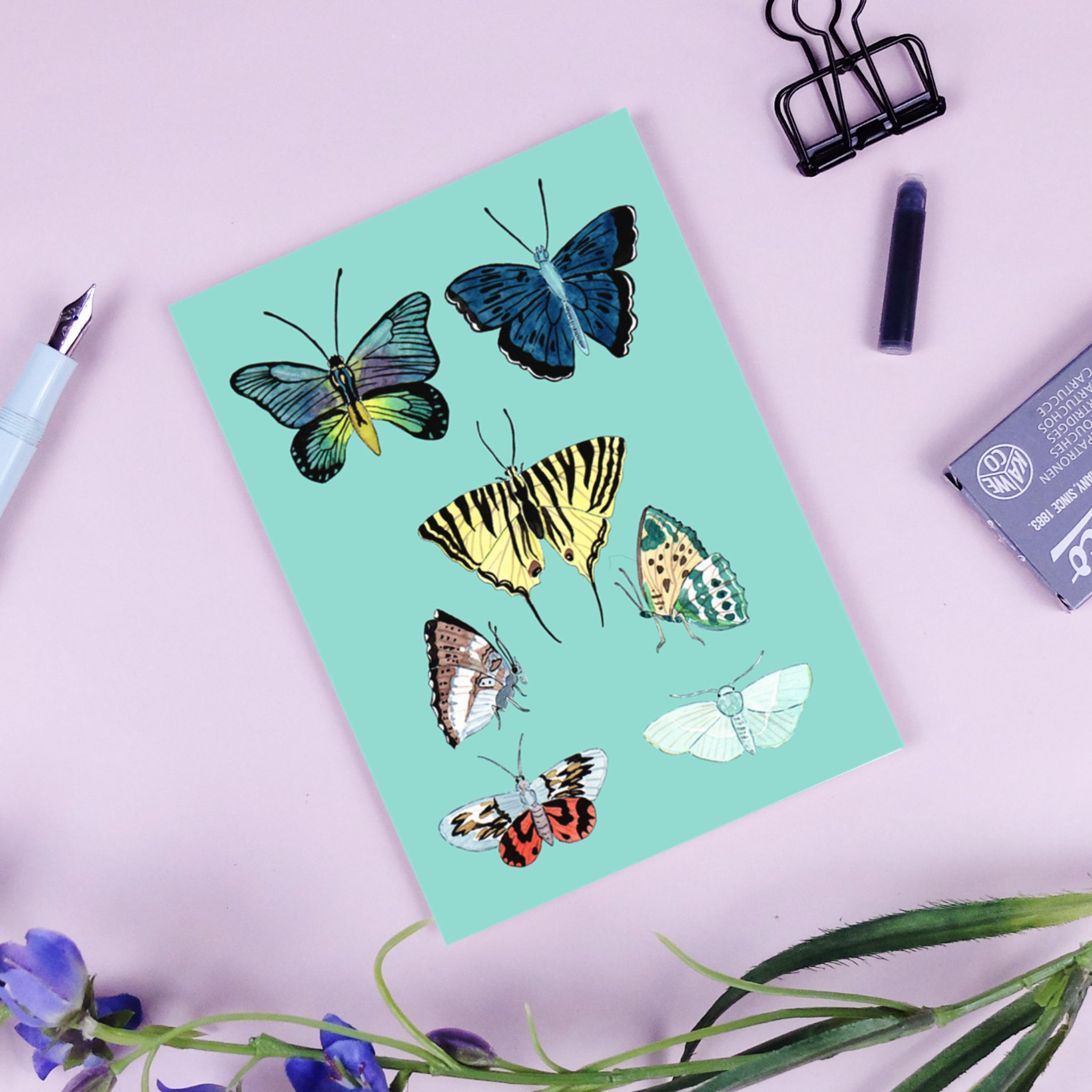 Illustration von acht bunten Schmetterlingen auf blauem Hintergrund, umgeben von Schreibwaren auf rosa Tisch.