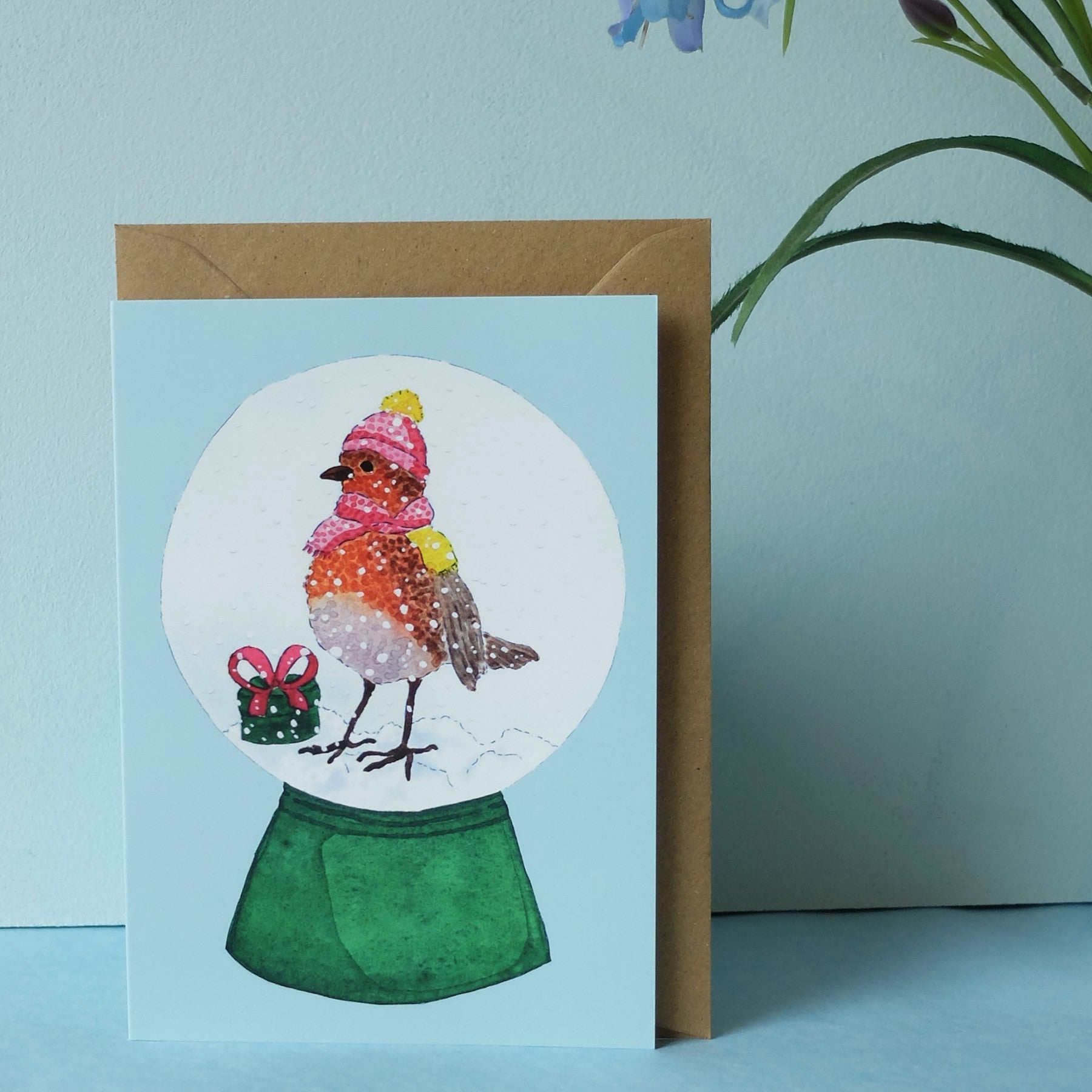 Eine Grußkarte mit einem Rotkehlchen mit rosa Mütze und Schal in einer Schneekugel, das neben einem kleinen grünen Geschenk mit roter Schleife steht. Die Karte befindet sich vor einem braunen Umschlag und einer hellblauen Wand mit einer Blume.