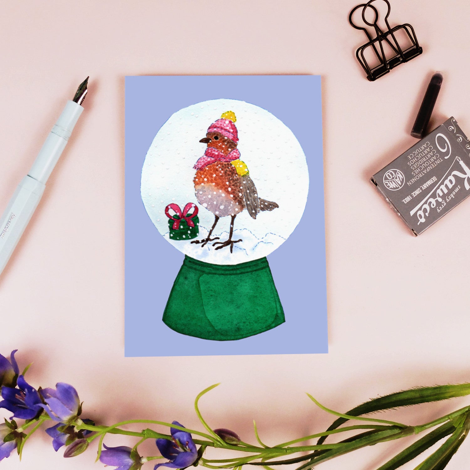 Eine Grußkarte mit einem Rotkehlchen in einer Schneekugel, das einen rosa Hut und Schal trägt, mit einem kleinen grünen Geschenk daneben. Die Karte befindet sich auf einer rosafarbenen Fläche mit einem Stift, einer Büroklammer, einem Radiergummi und lila Blumen in der Nähe.