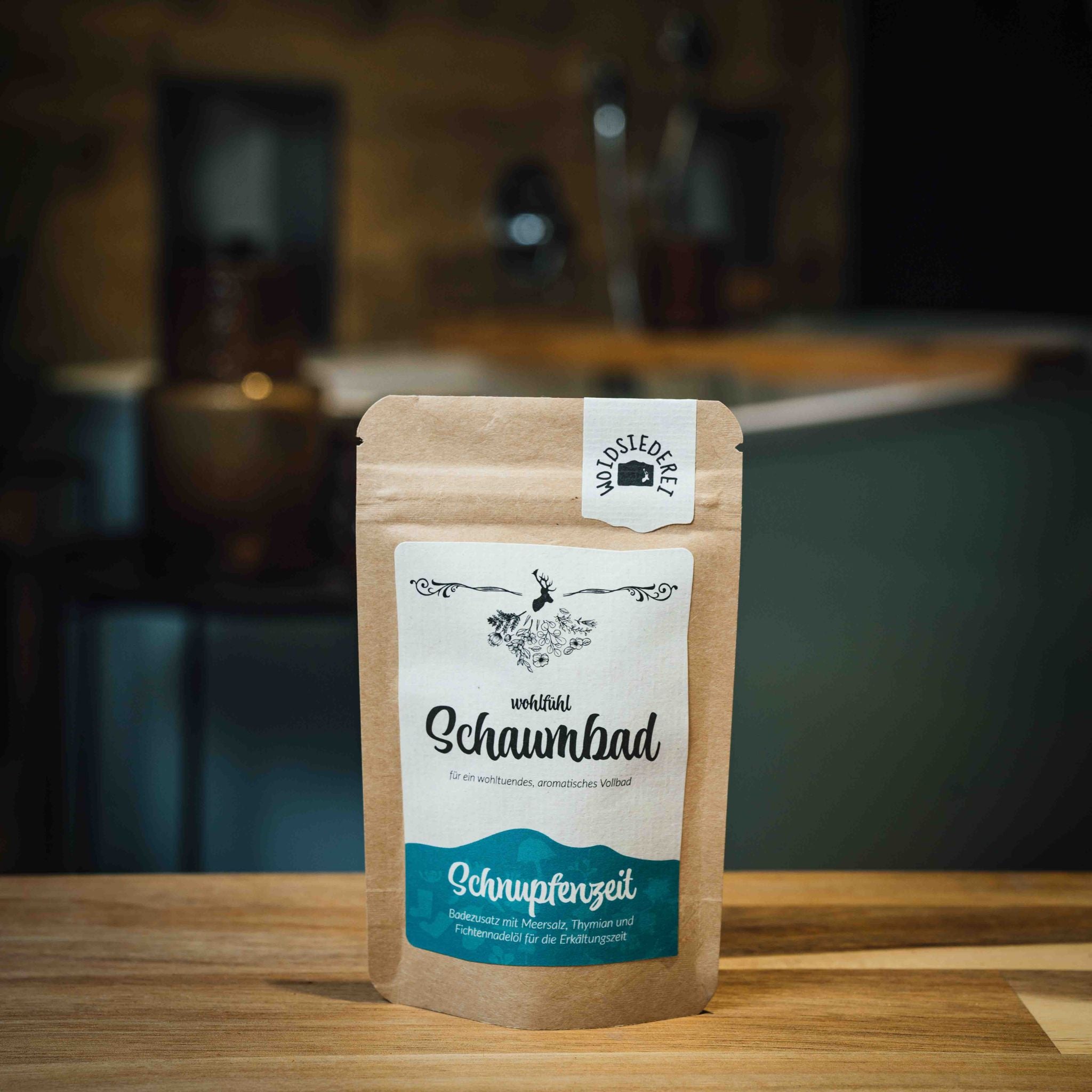 Braune Packung mit Aufschrift "Schaumbad" und "Schnupfenzeit" auf einem Holztisch, unscharfer Hintergrund.