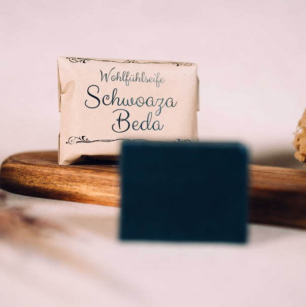 Packung der "Wohlfühlseife Schwoaza Beda" und ein dunkles Seifenstück auf einem Holzbrett.
