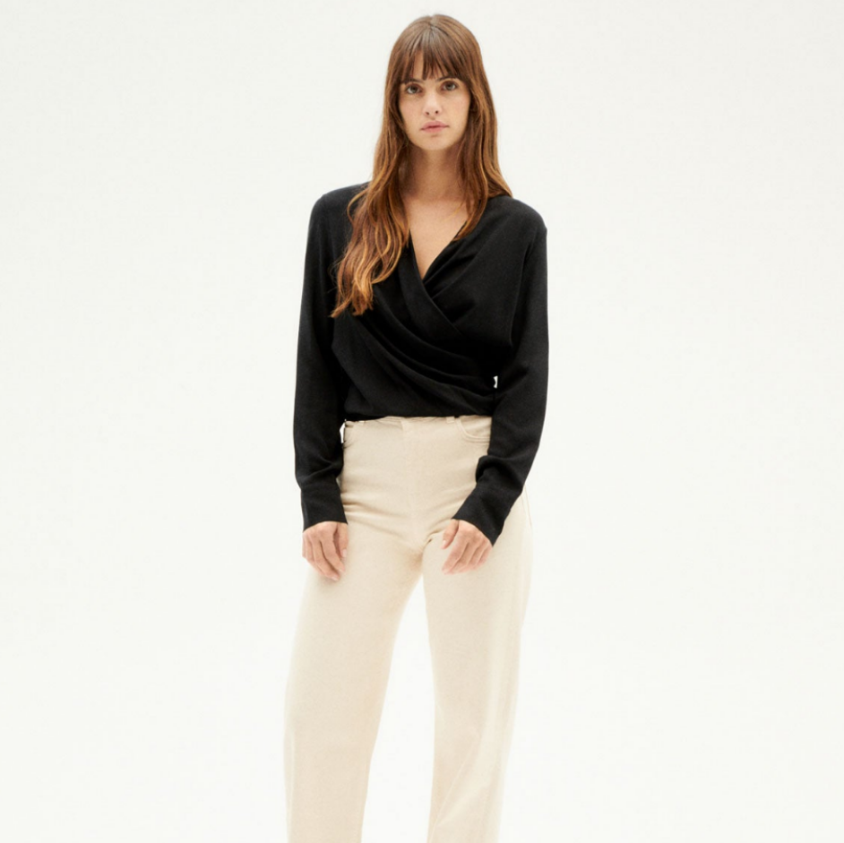 Eine Frau mit langen braunen Haaren steht vor einem schlichten weißen Hintergrund und trägt ein schwarzes langärmeliges Wickeloberteil und eine beigefarbene Hose mit hoher Taille und weitem Bein.