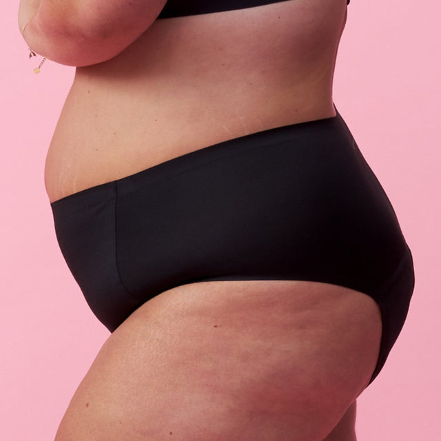 Eine Person mit heller Haut, die schwarze Unterwäsche und einen schwarzen BH trägt, posiert im Profil vor einem rosa Hintergrund. Ihr Arm ist angewinkelt und der Fokus liegt auf ihrem Mittelteil und ihren Hüften.