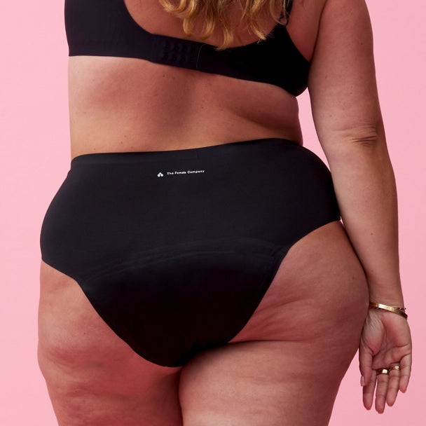 Eine Frau mit gewelltem blondem Haar trägt einen schwarzen BH und schwarze Unterwäsche mit hoher Taille und steht mit dem Rücken zur Kamera vor einem rosa Hintergrund.