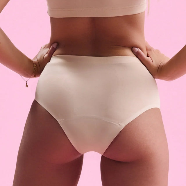 Eine Frau steht mit den Händen in den Hüften, trägt einen beigen BH und hochtaillierte Unterwäsche vor einem einfarbig rosa Hintergrund.