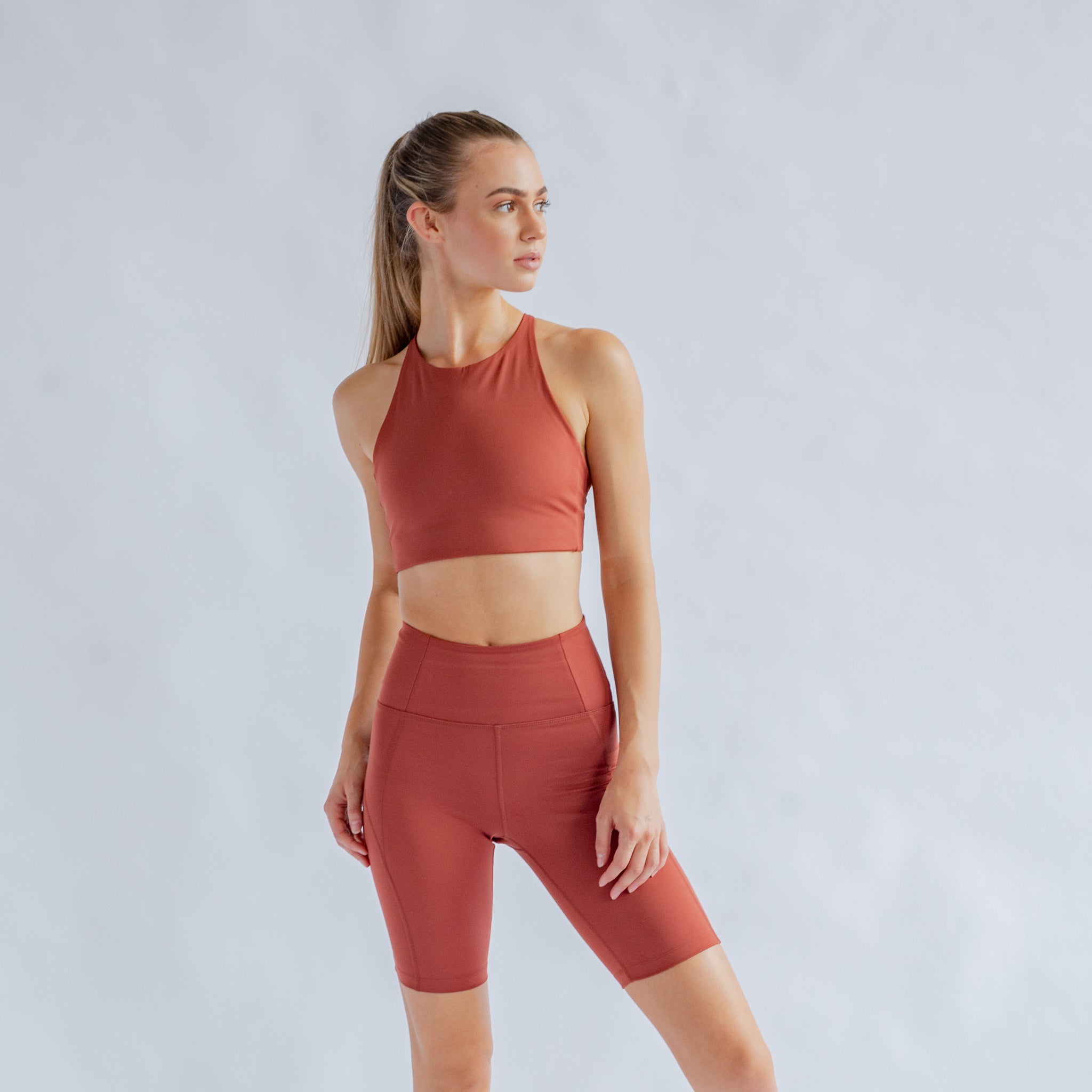 Frau in rostfarbenem Sportset blickt zur Seite, vor grauem Hintergrund.