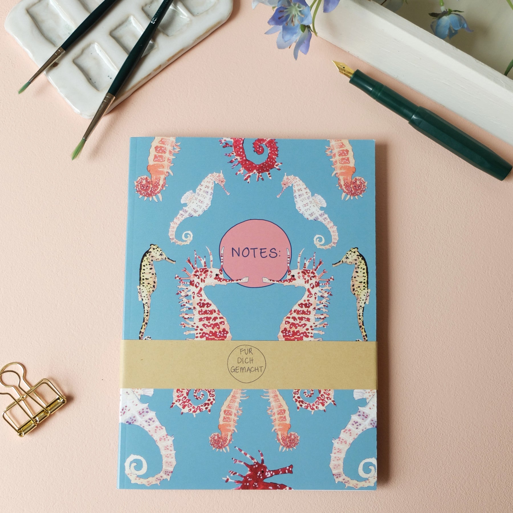 Ein Notizbuch mit Seepferdchen- und Meeresdesign auf blauem Hintergrund, umgeben von Malzubehör und Blumen.