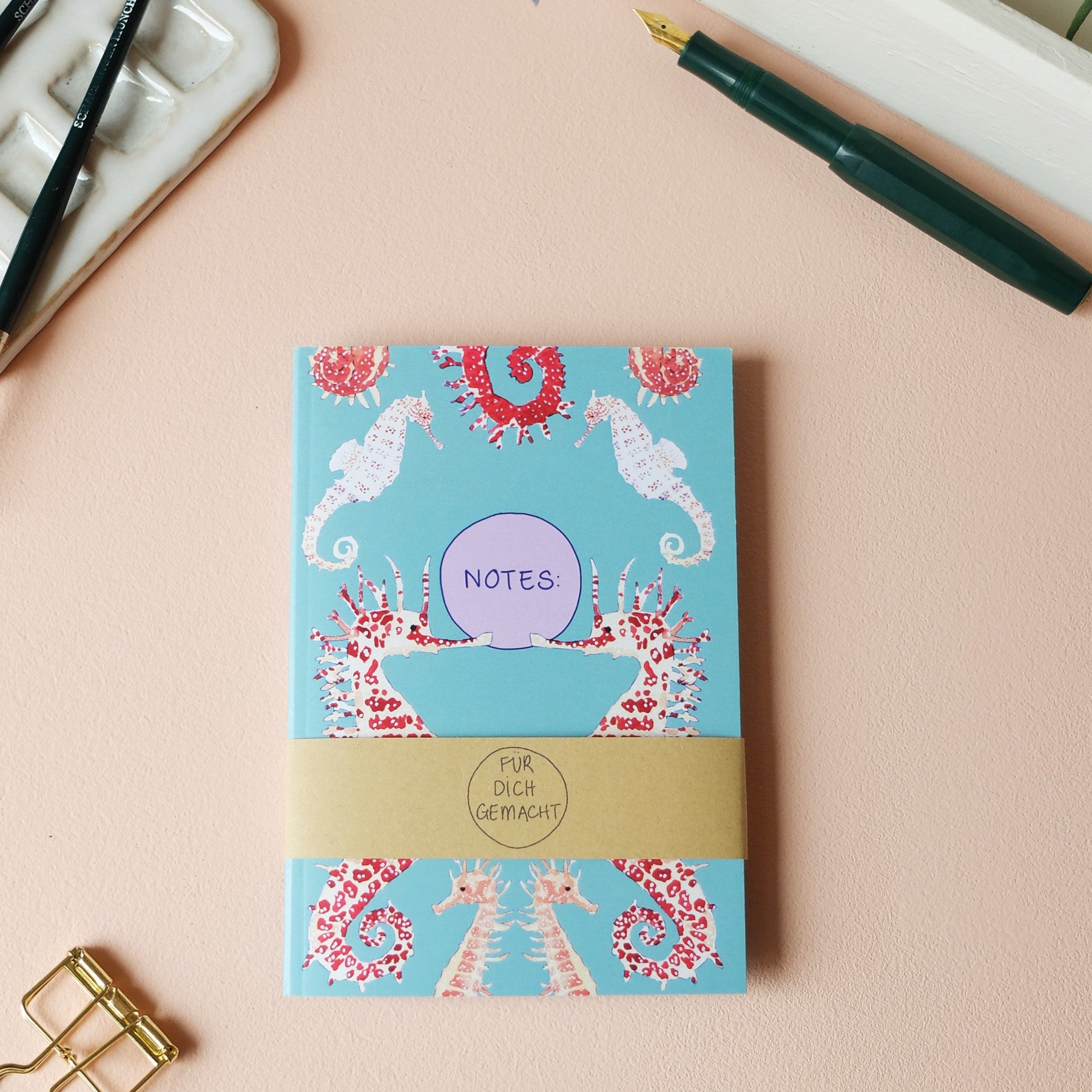 Notizbuch mit Seepferdchen-Muster auf blauem Hintergrund, umgeben von Stiften und Büroklammer auf einem rosa Tisch.