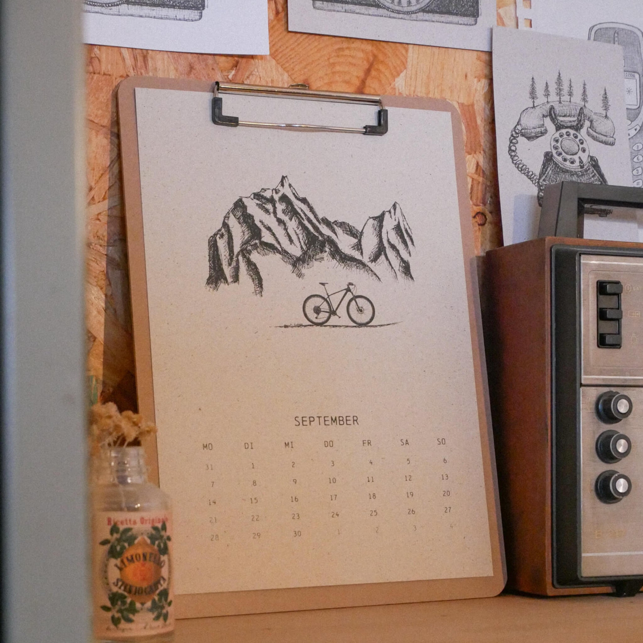 Auf einem Klemmbrett liegt ein September-Kalenderblatt mit einer Illustration eines Fahrrads und von Bergen. Es steht auf einem Regal neben einem alten Radio, einem kleinen Krug und verschiedenen Kunstdrucken, die an eine Holzwand gepinnt sind.