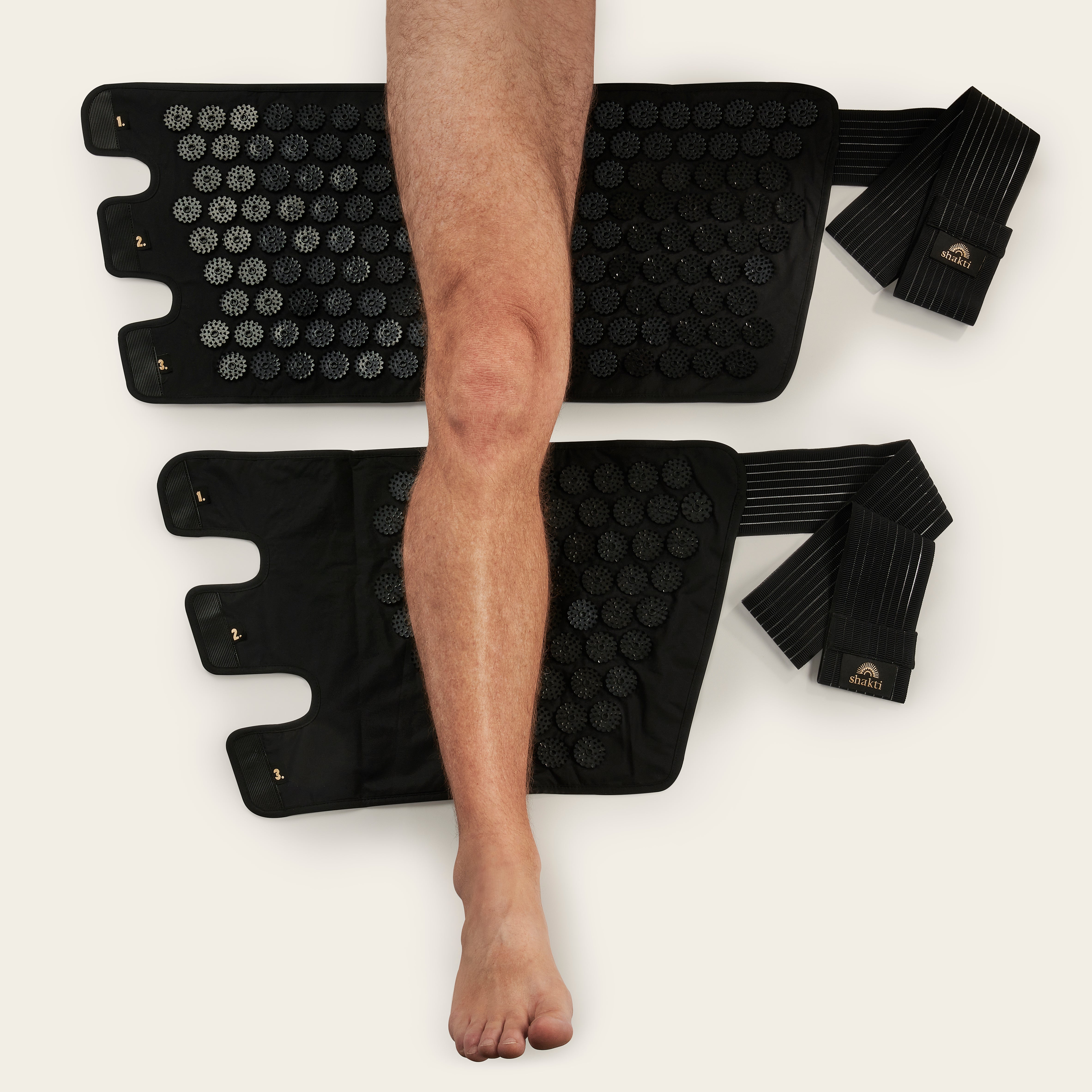Das nackte Bein einer Person ruht auf zwei schwarzen Akupressur-Matten und zwei elastischen Bändern mit Klettverschluss und Logo, die auf einem hellen Hintergrund ausgelegt sind.